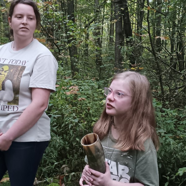 Two young girls standing in a forest surrounded by green foliage. One girl is holding a large seed pod or shell and the other girl is wearing glasses and looking at her.
