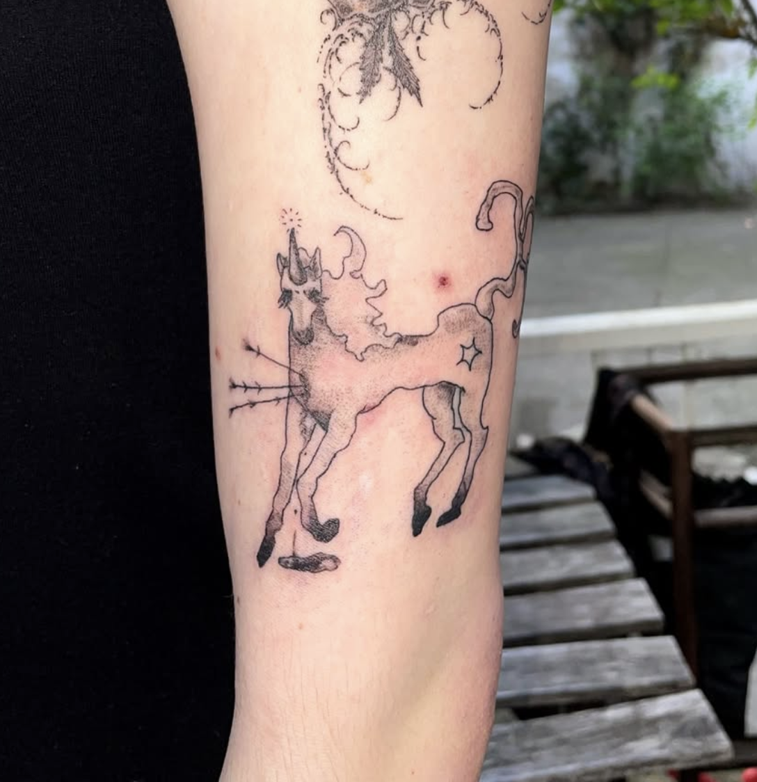 Tattoo eines Einhorns in Schwarz auf dem Oberarm einer Person, im Hintergrund sind Wolken und Sterne sichtbar.