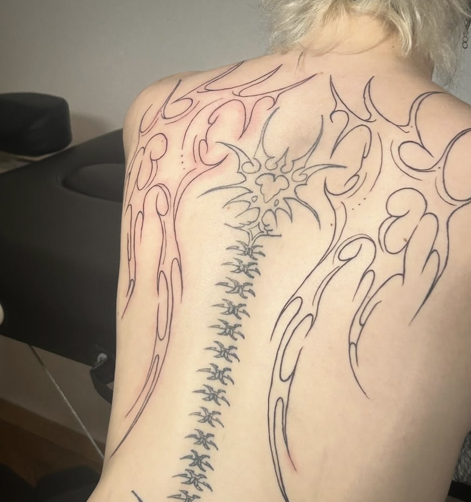 Eine Person mit einem tribalähnlichen Tattoo-Design, das von der Schulter bis zur Wirbelsäule reicht, auf der Haut. Das Tattoo besteht aus schwarzen Linien und Mustern.
