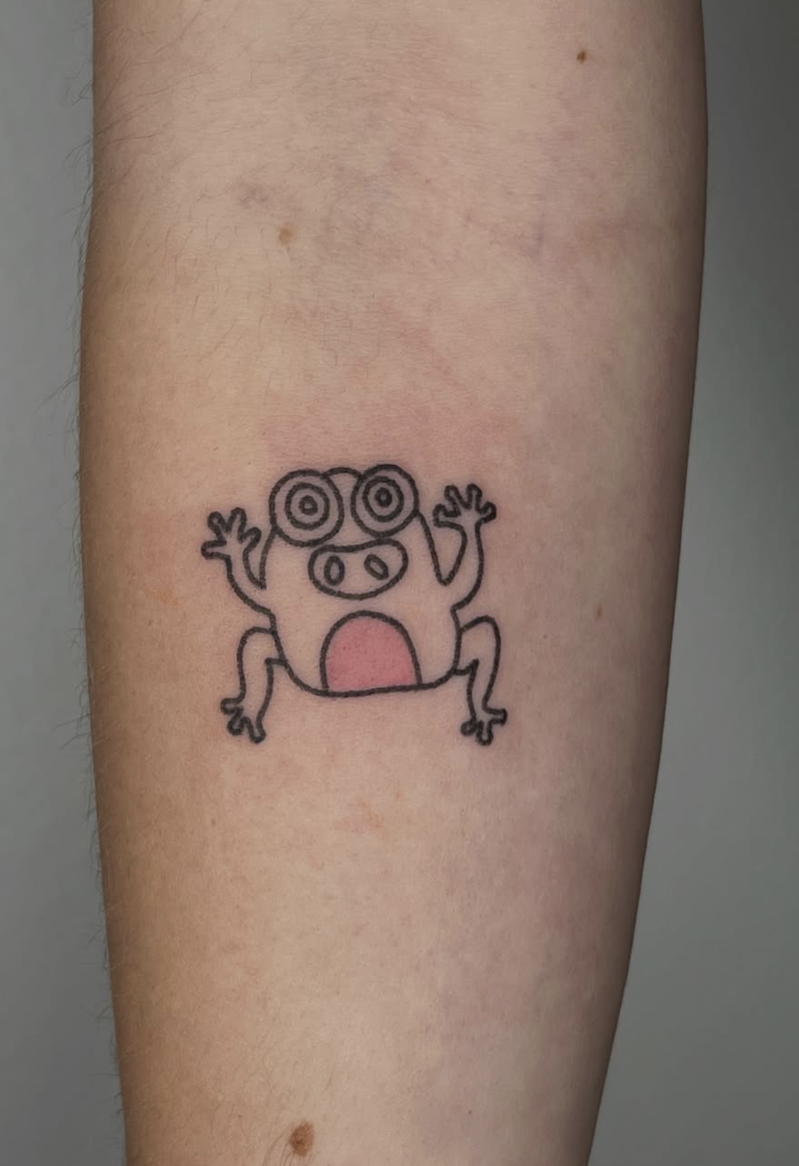 Tattoo eines cartoonartigen Frosches mit großen, spiralförmigen Augen, offenem Mund und gespreizten Armen, auf einem menschlichen Arm.