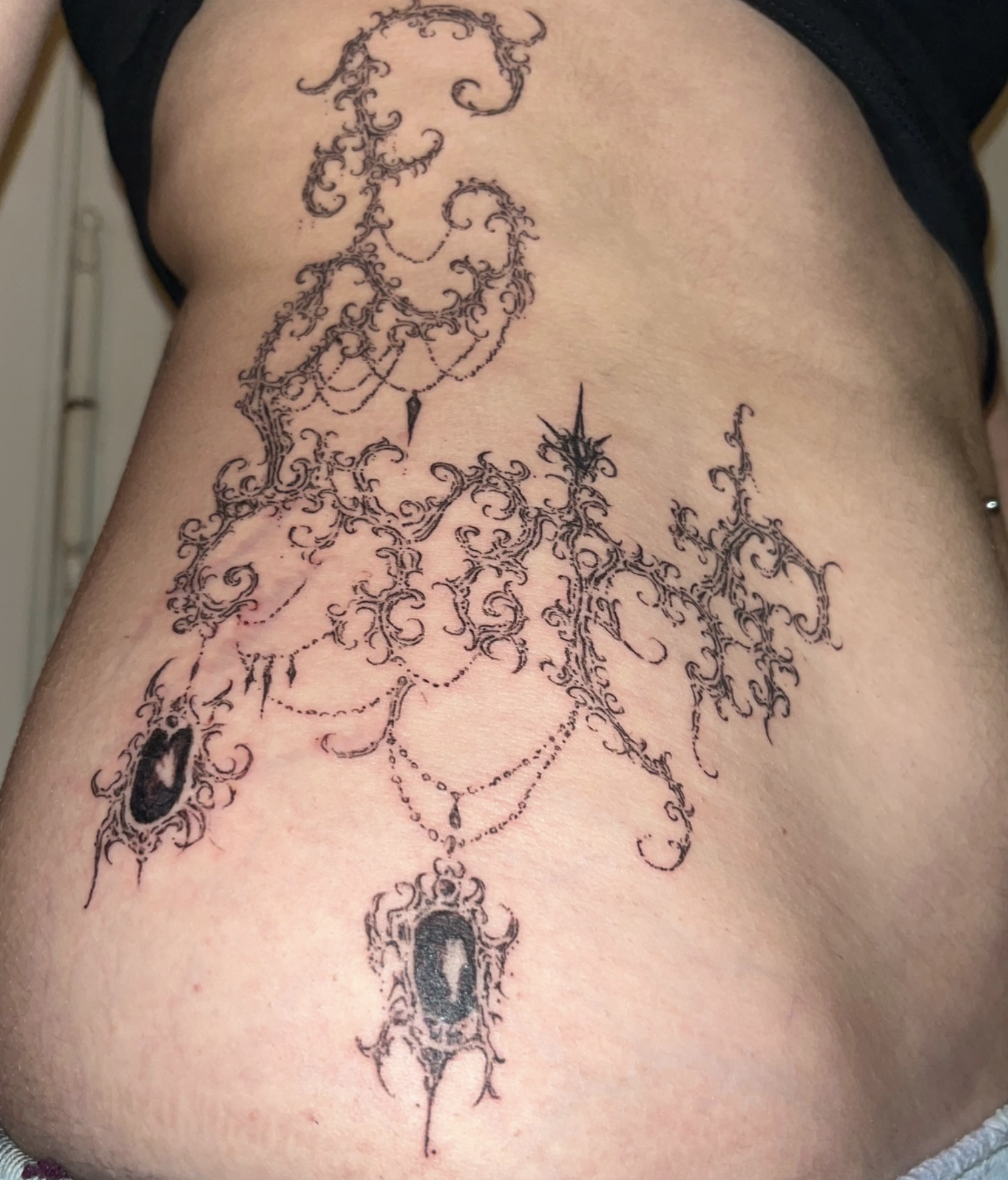 Tattoo auf der Seite, mit schwarzen, filigranen, barocken Ornamenten, einem Spiegelrahmen und einer Waage.