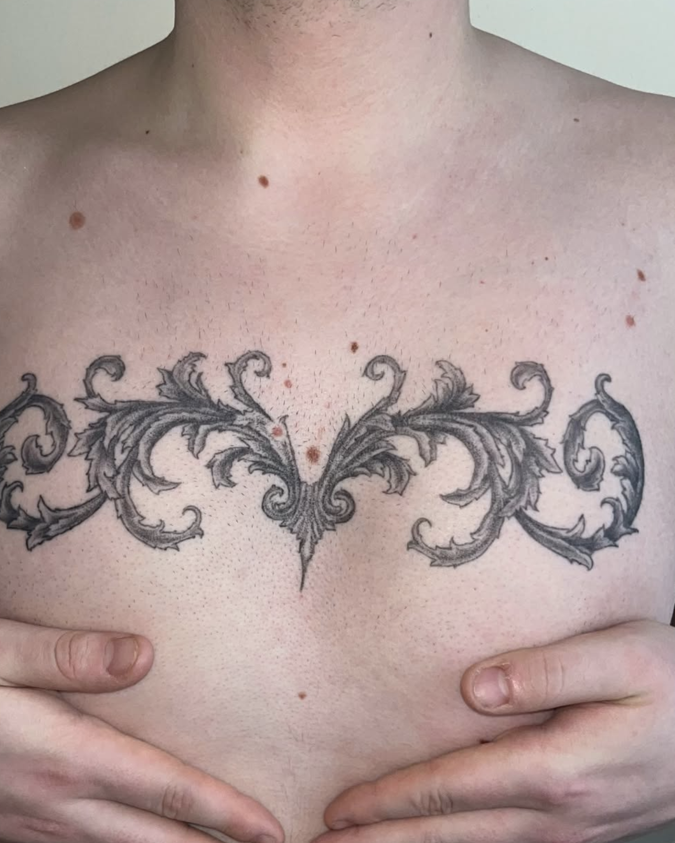 Nahaufnahme eines männlichen Oberkörpers mit einer großen schwarzen Tattoo-Bekrönung auf der Brust, Hände halten den unteren Brustbereich.