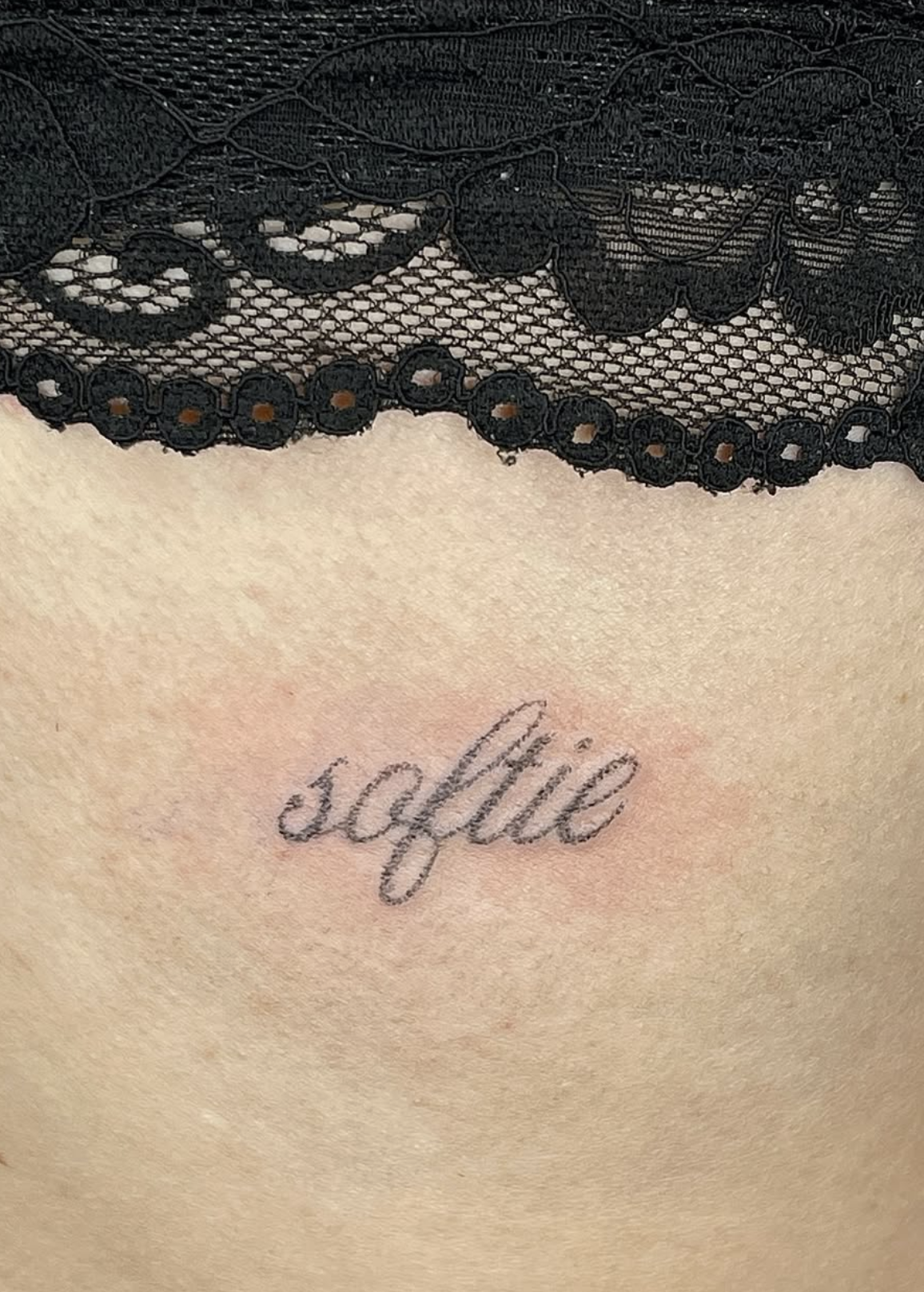 Nahaufnahme eines Tattoos mit dem Wort 'sophie' in Schreibschrift, darunter sichtbarer schwarzer Spitzenstoff und Haut.