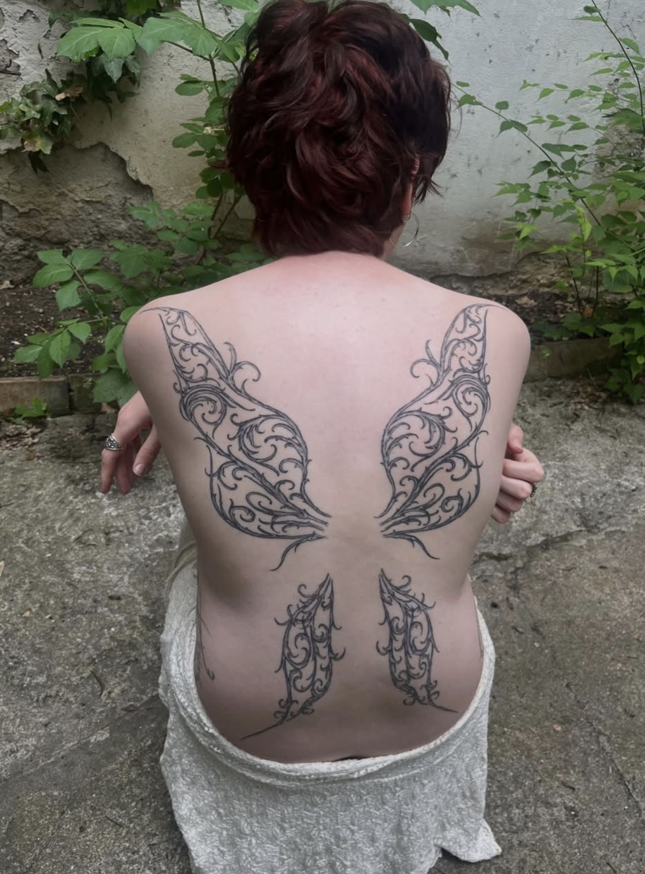 Frau mit Rückenansicht, tätowierte Flügel auf dem Rücken, rote Haare, sitzt im Freien vor einer Steinmauer mit Pflanzen.