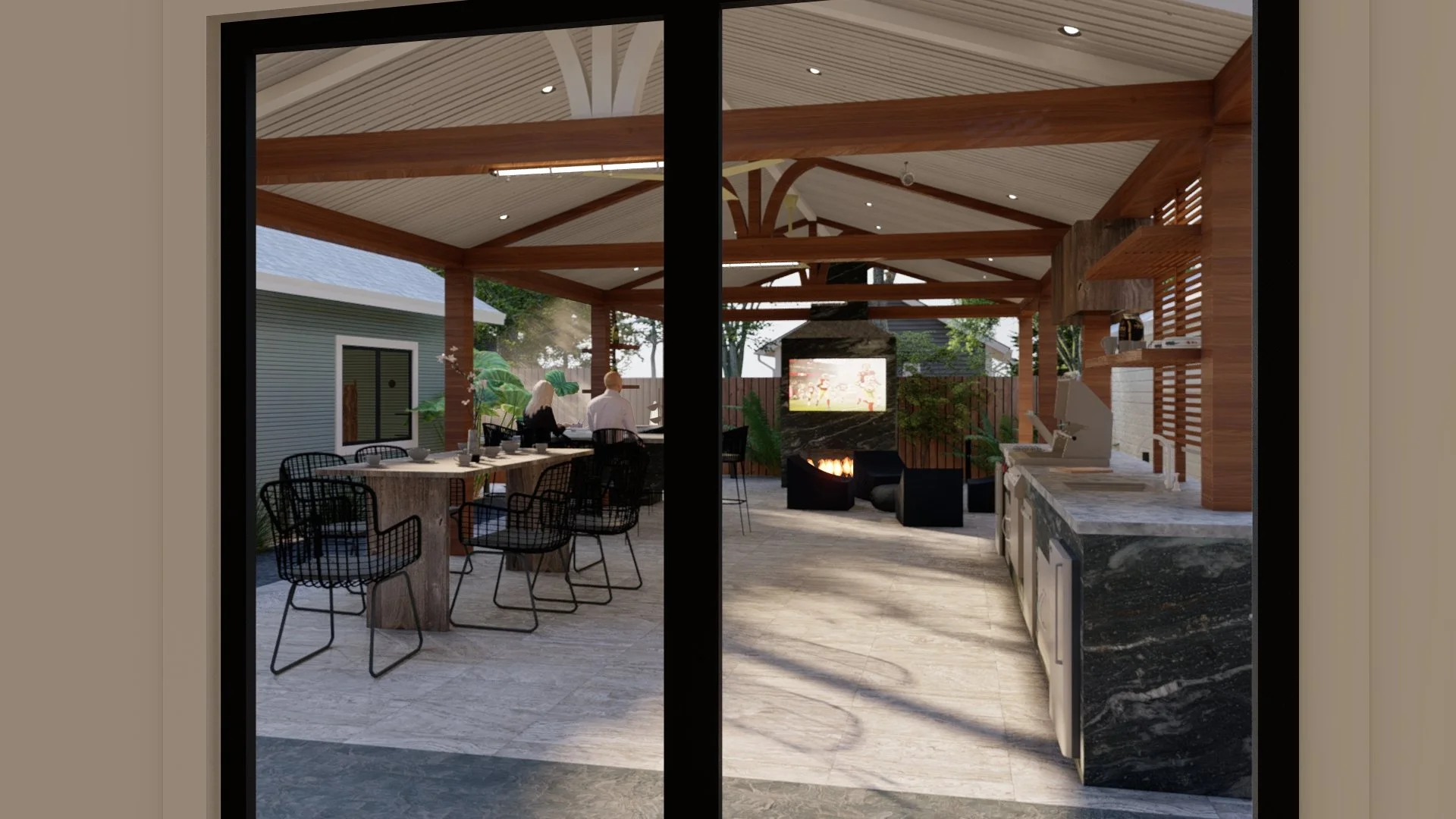 View through a large window into a modern outdoor living space with a covered patio featuring a dining area, a fireplace with a television above, and a kitchen countertop with a coffee machine. There are two people standing near a bar counter, lush greenery, and a fence in the background.