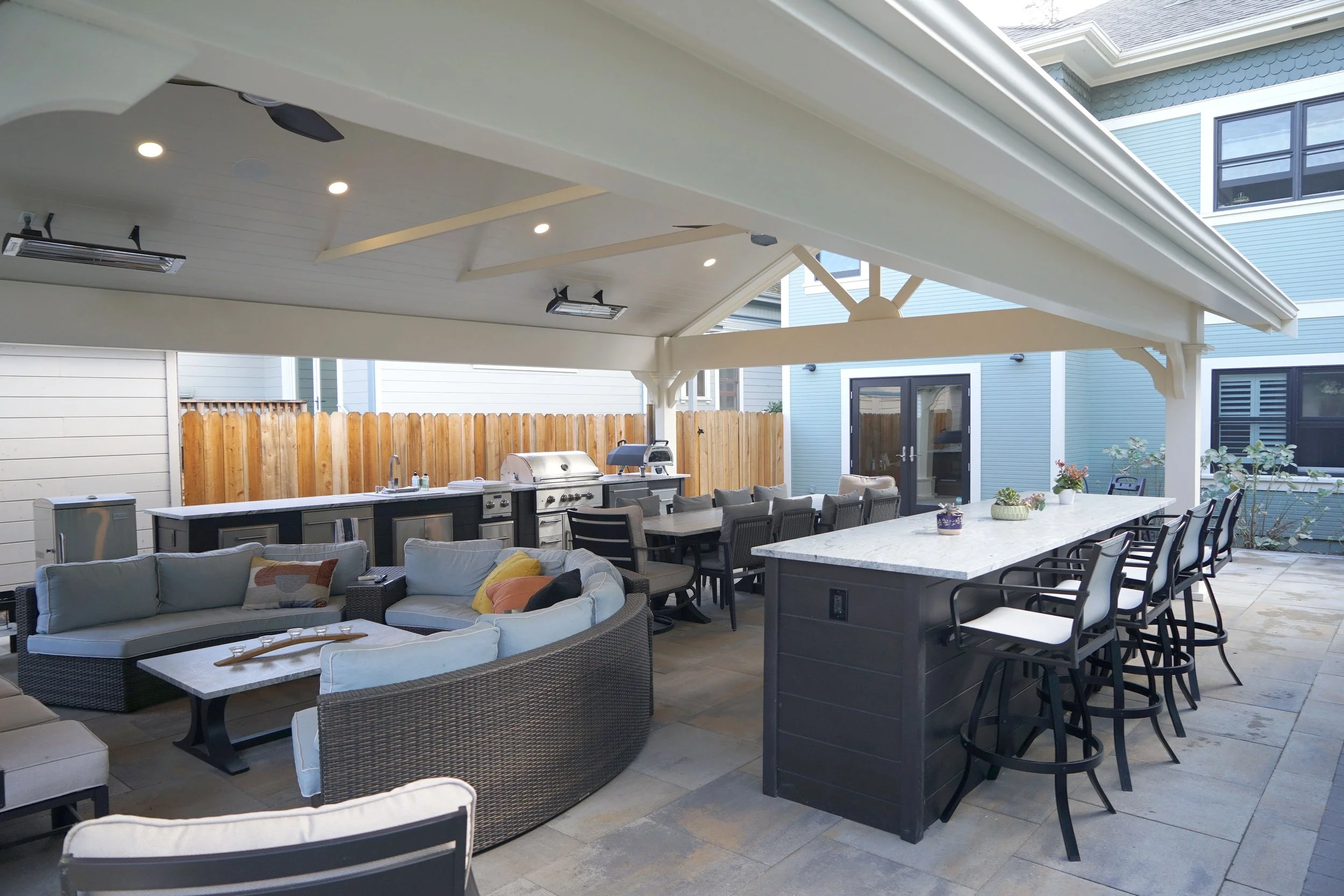 Covered outdoor patio with seating, a kitchen area, and a bar with high chairs in a backyard setting.