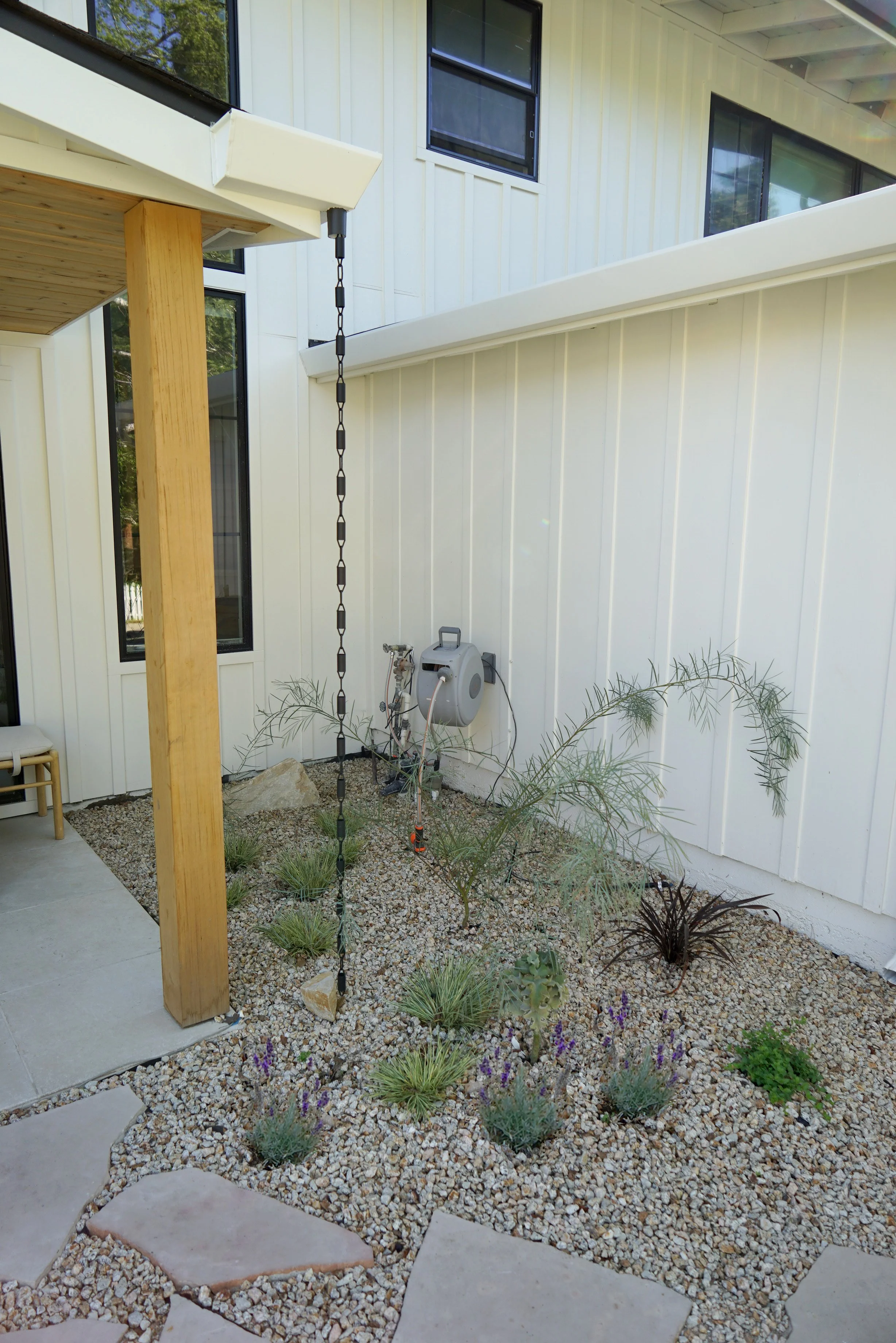 A small backyard garden with desert plants, gravel, a hose reel, and a white modern house with large windows.