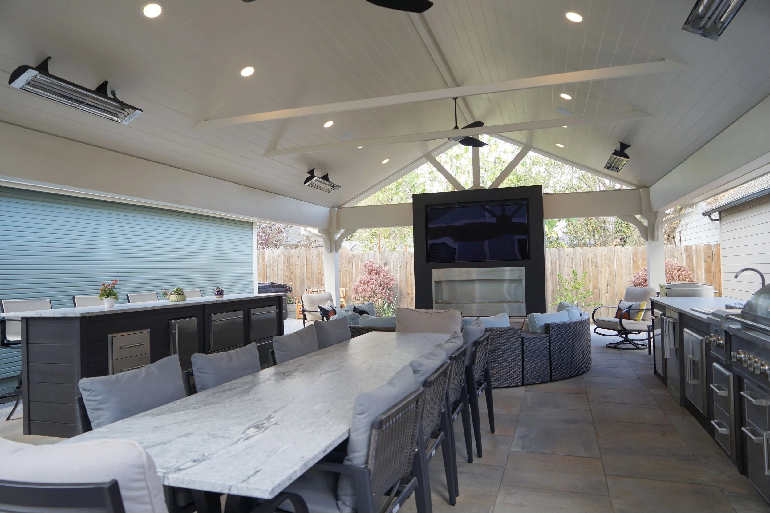 Covered outdoor patio with a long dining table and chairs, a kitchen area with appliances, a built-in fireplace, a large television, outdoor seating, ceiling fans, and landscaping visible through open sides.