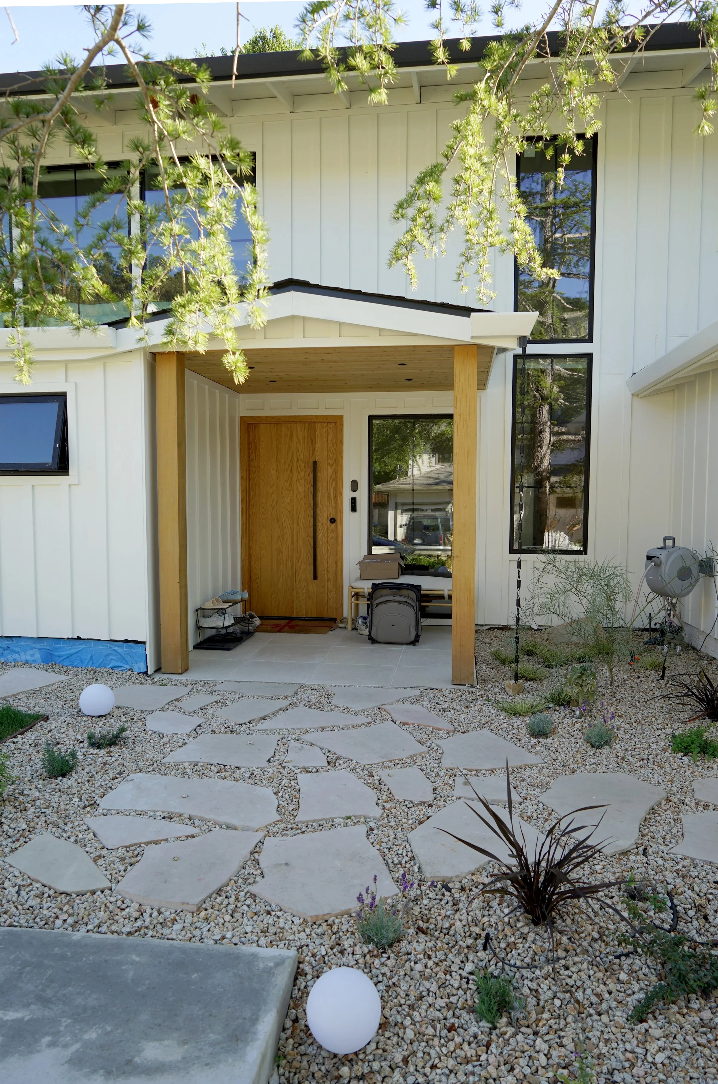 Front entrance of a modern house with a wooden door, small porch, large black-framed windows, and a stone pathway surrounded by gravel and drought-tolerant plants.