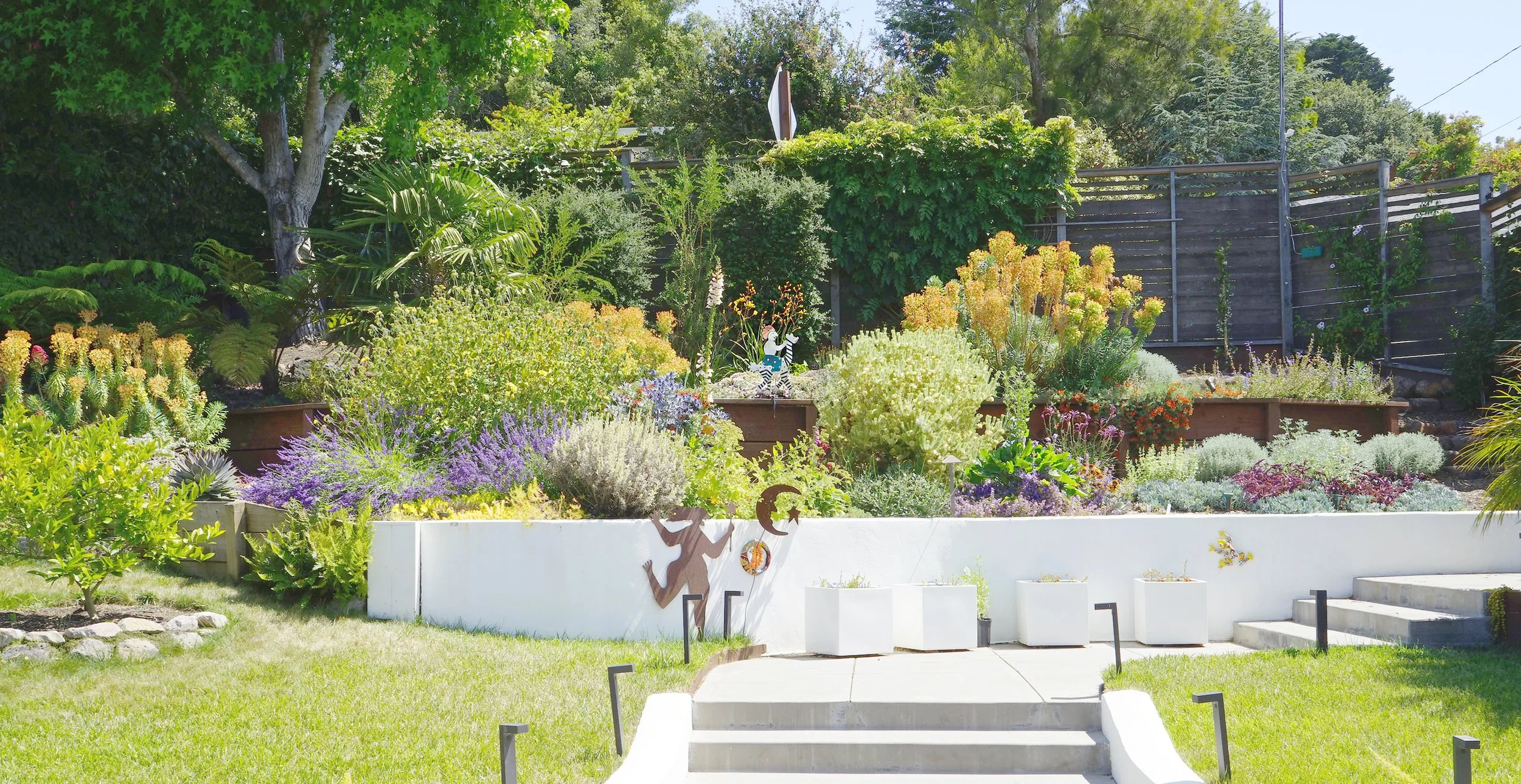 A tiered garden with vibrant flowering plants and shrubs, a white painted wall with decorative metal art, and a grassy yard with stairs leading to the garden, surrounded by trees and a weathered wooden fence.