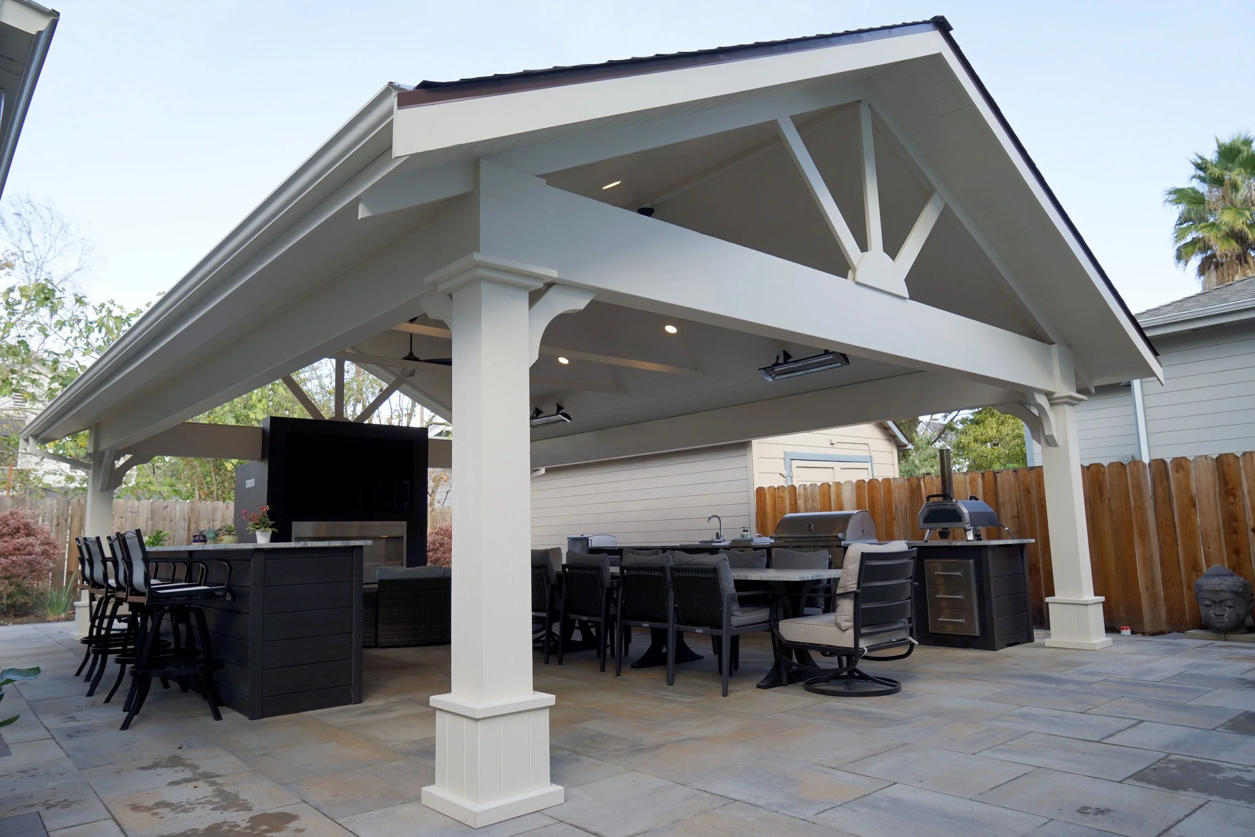 Outdoor covered patio area with black and beige furniture, a television, built-in grill, and wooden fencing.