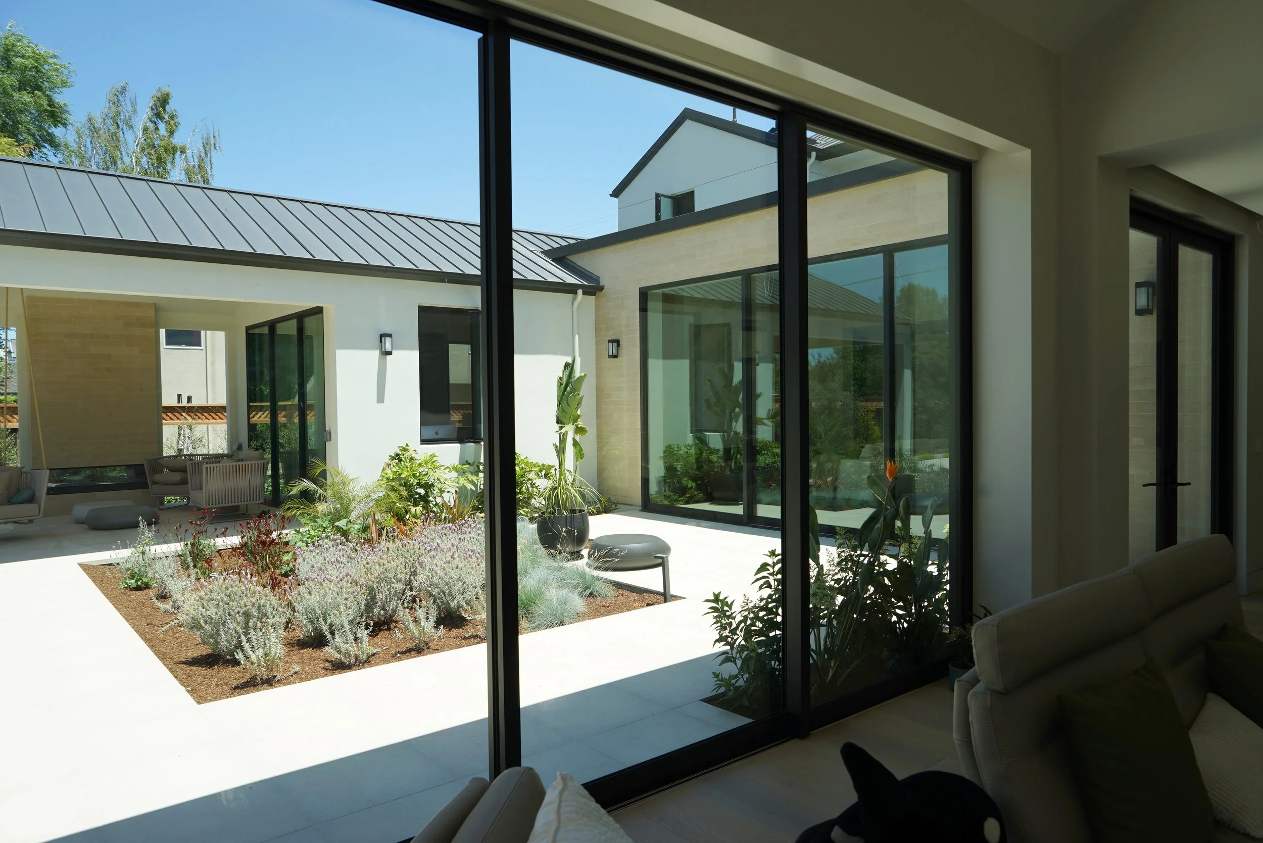 View of a modern backyard patio with plants and outdoor seating, seen through large sliding glass doors from the interior of a house.