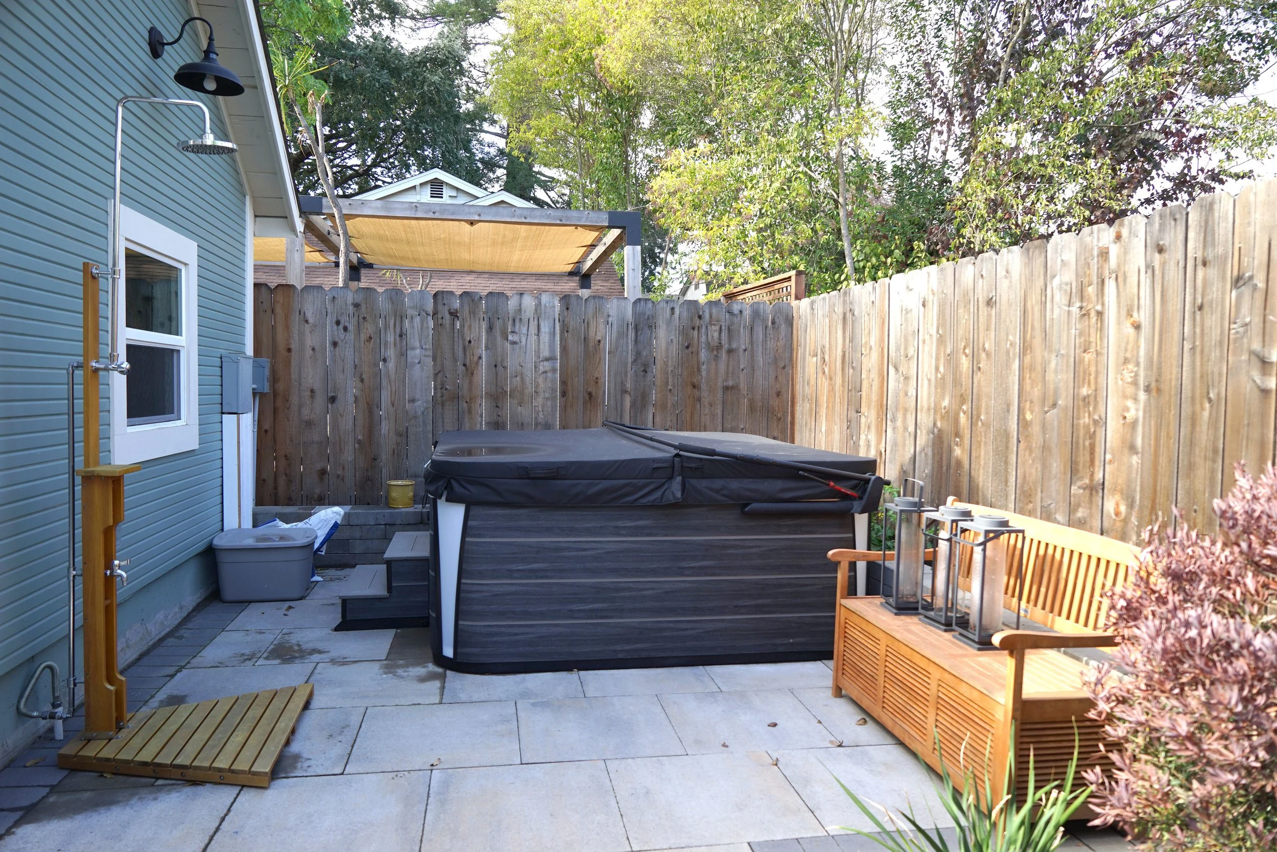 Backyard patio with a hot tub covered with a black cover, a wooden bench with lanterns on it, a outdoor shower, and a wooden fence. There are trees and a structure with a beige canopy in the background.