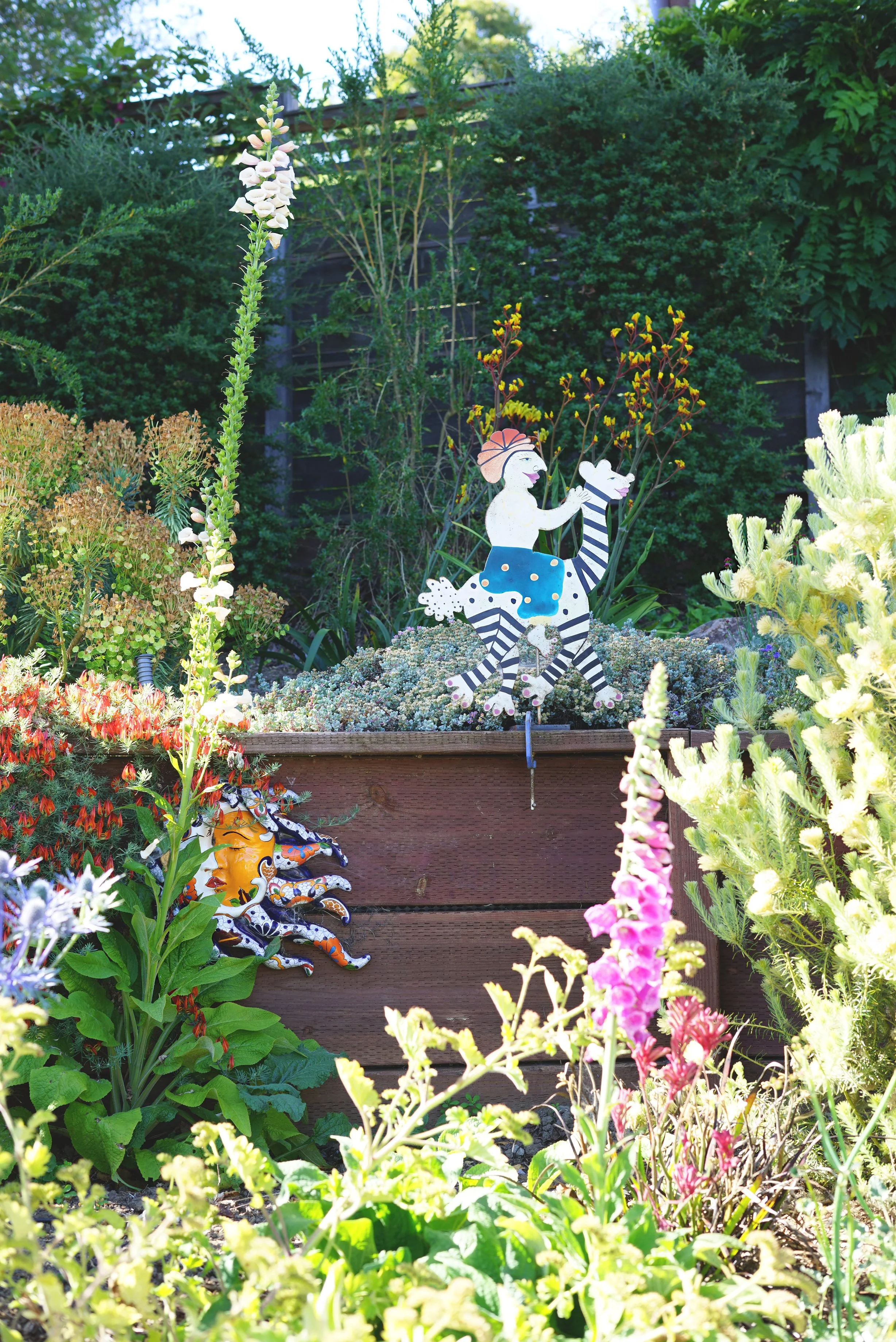 Colorful garden with decorative metal sculptures of a woman riding a zebra and a sun face among vibrant flowers and greenery.