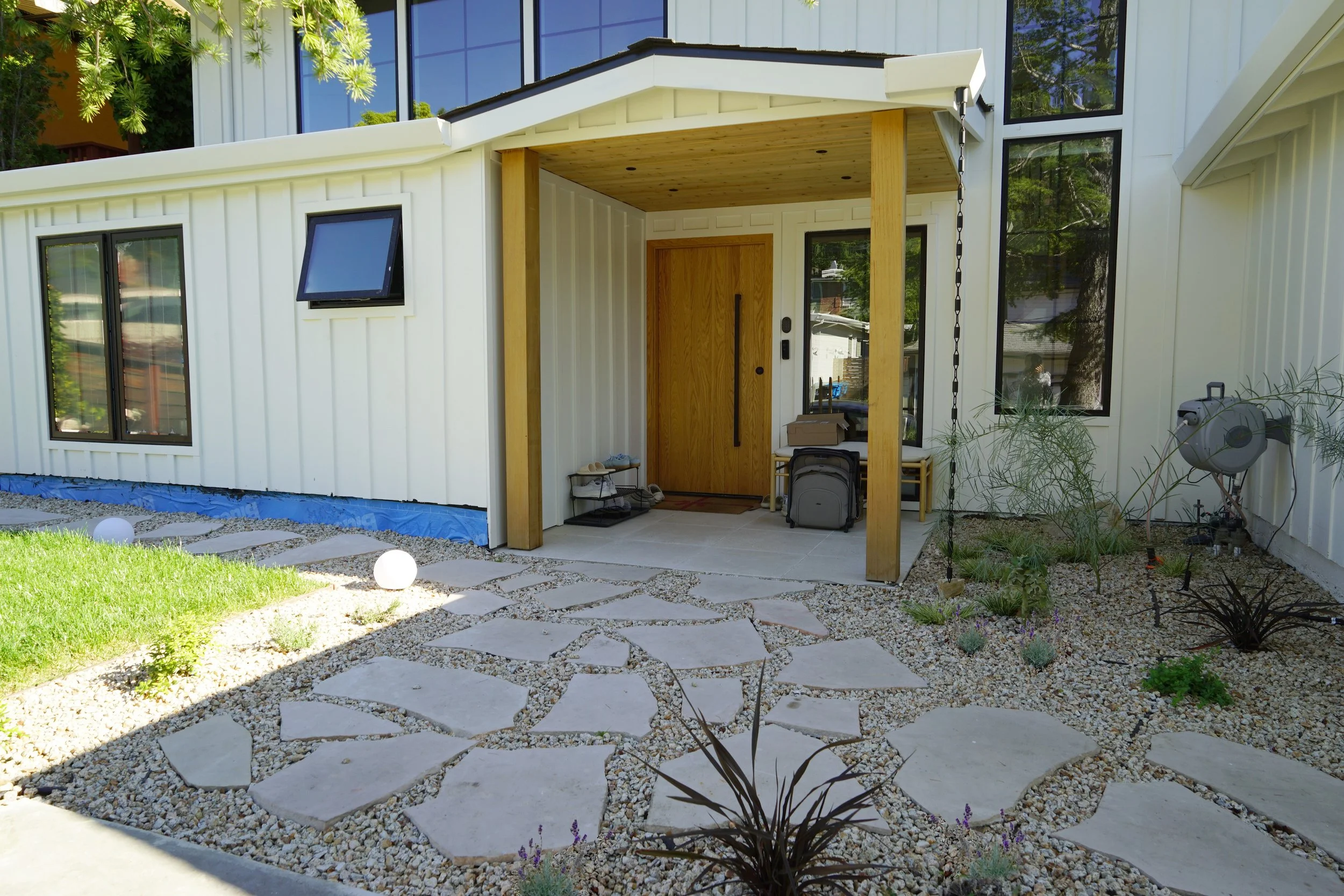 Modern house exterior with a small covered porch, wooden front door, multiple windows, landscaped yard with stepping stones, small plants, and outdoor lighting.