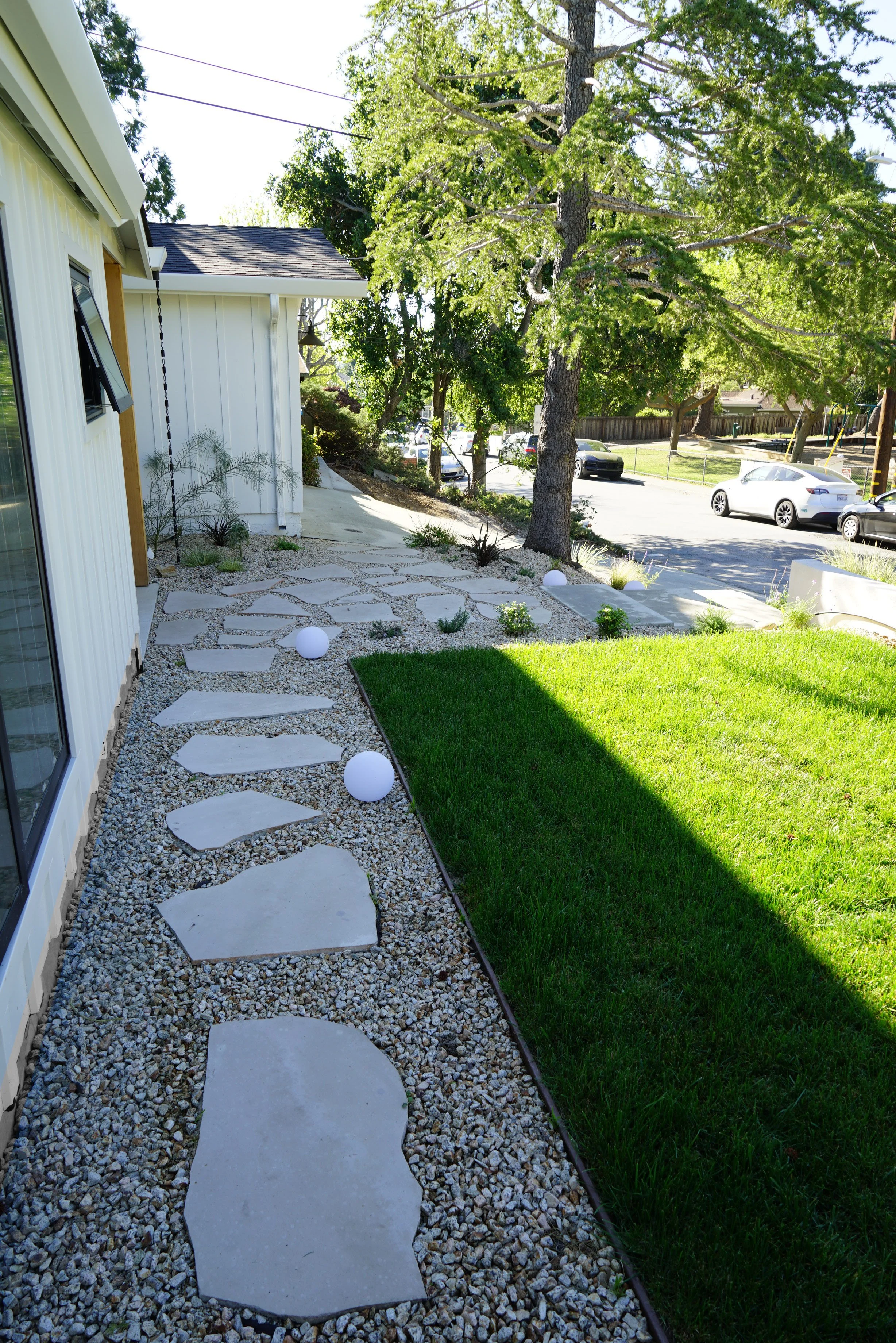 A modern backyard with stepped stone pathway, gravel ground cover, small bushes, perennials, a tree, coiled garden hoses, illuminated by white spherical garden lights, with a grassy lawn and neighboring street with parked cars.