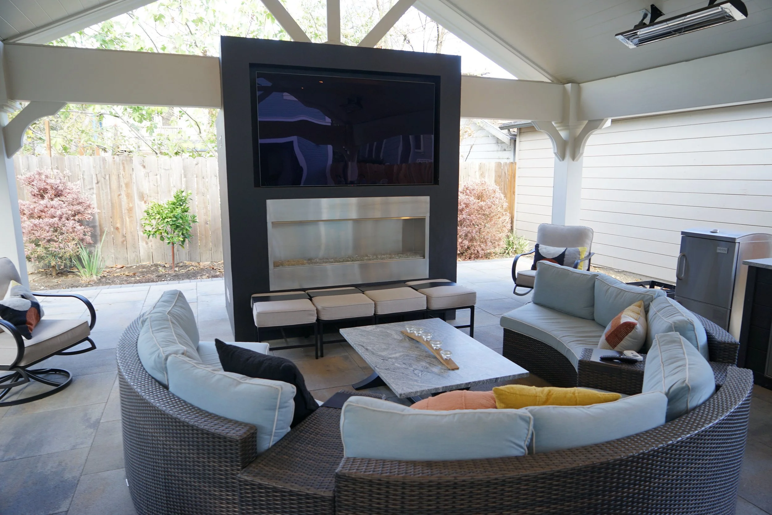 Outdoor covered patio with a curved wicker sectional sofa with light cushions, a marble coffee table with candles, a white cushioned armchair, a smaller cushioned chair, a fireplace built into a black wall with a mounted television above, and a small silver refrigerator on the right, with outdoor plants and a wooden fence in the background.