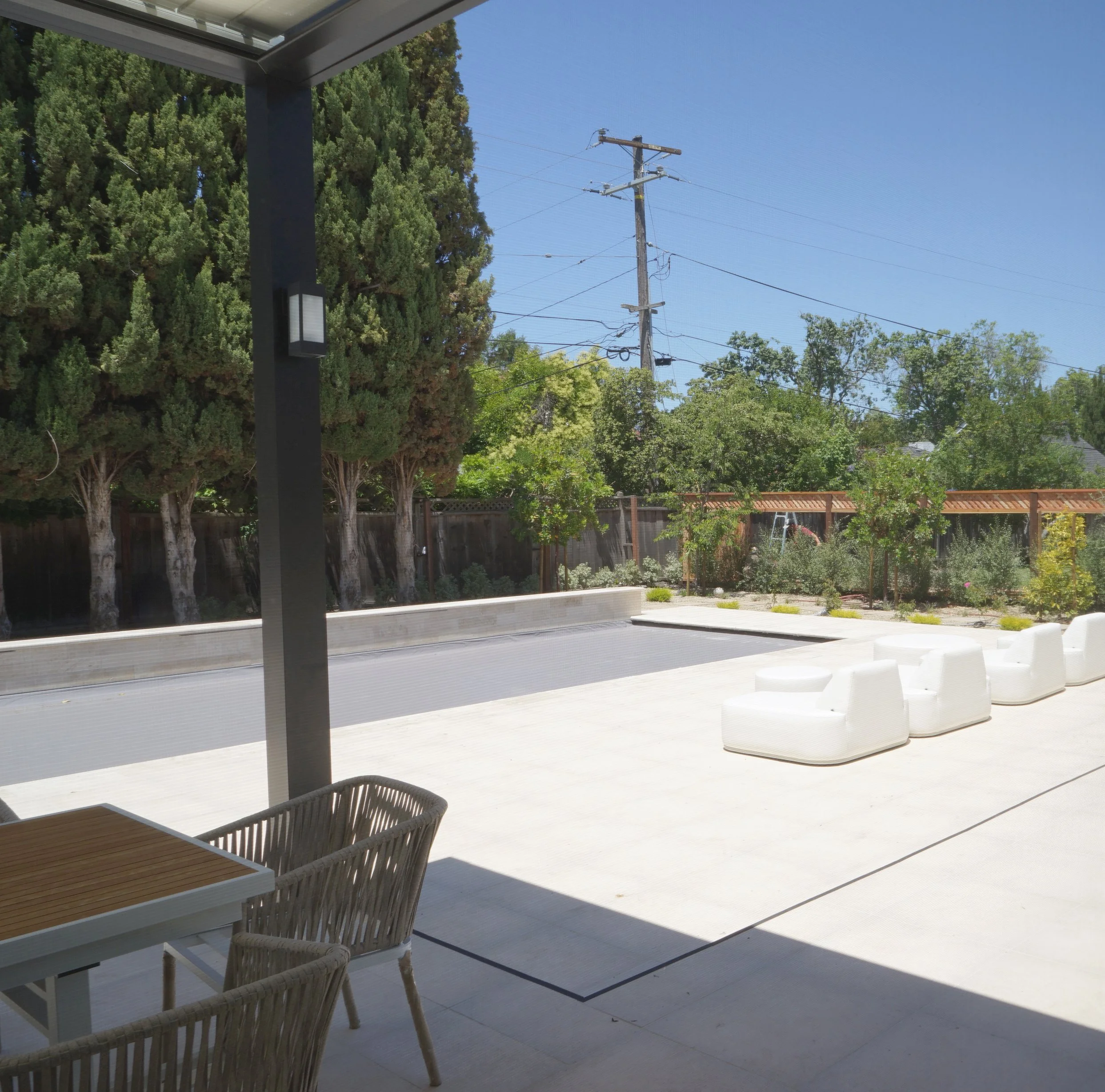 A backyard patio with white outdoor chairs, a wooden table with wicker chairs, tall trees, a wooden fence, and utility pole with power lines under a blue sky.