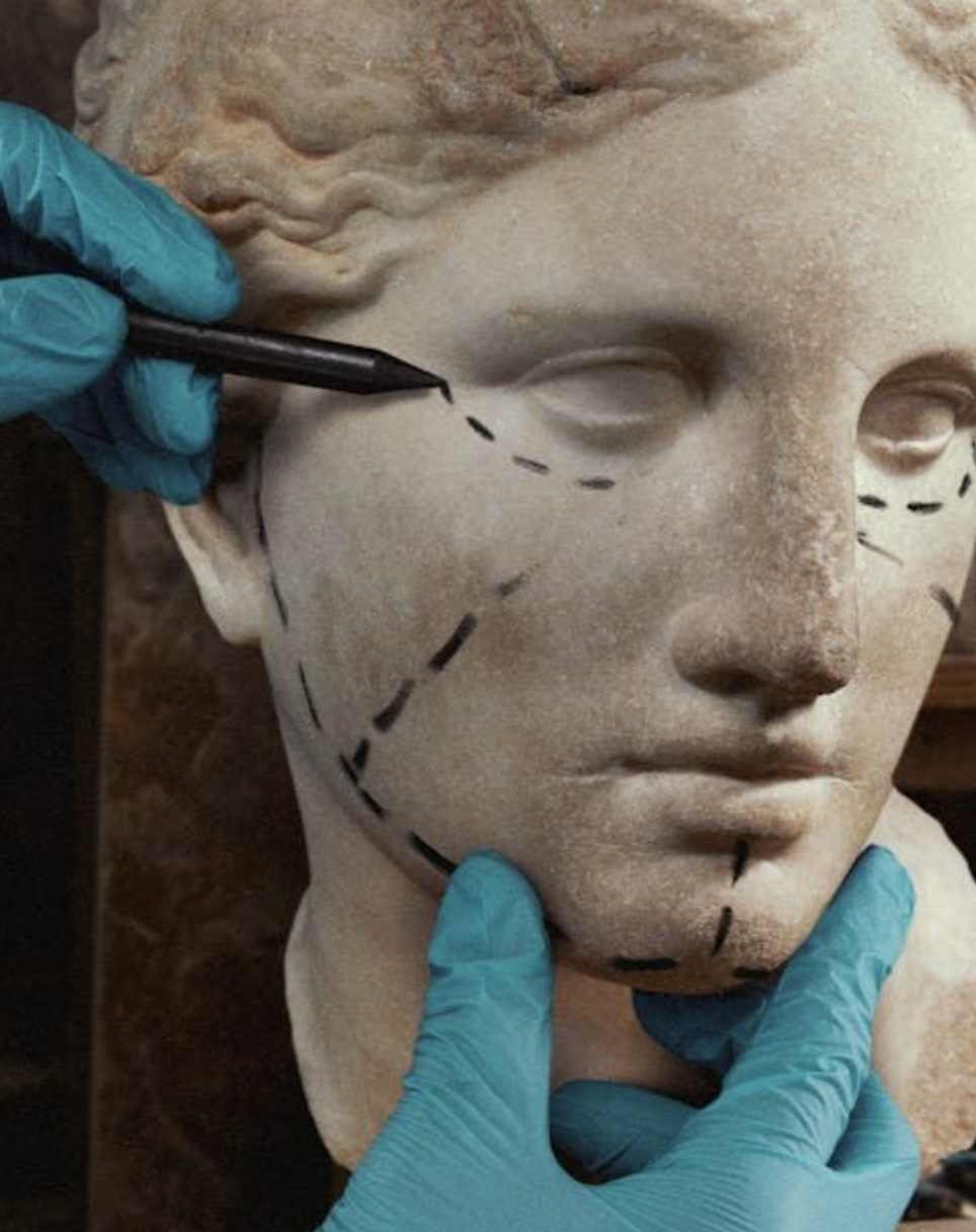 Restorer's hands in blue gloves marking a marble bust of a woman's face with a black marker, indicating planned restorations or repair areas.