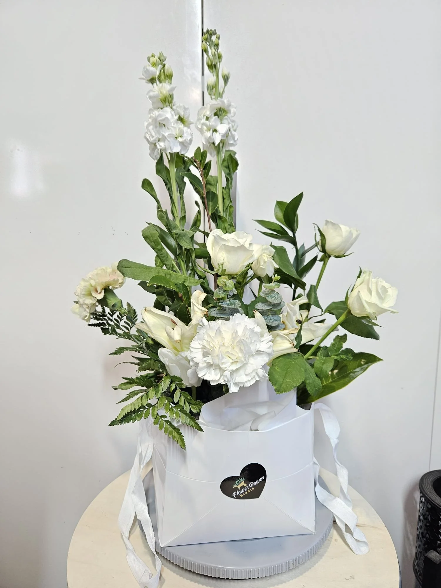 White flower arrangement with roses, carnations, and greenery in a white gift bag on a table.