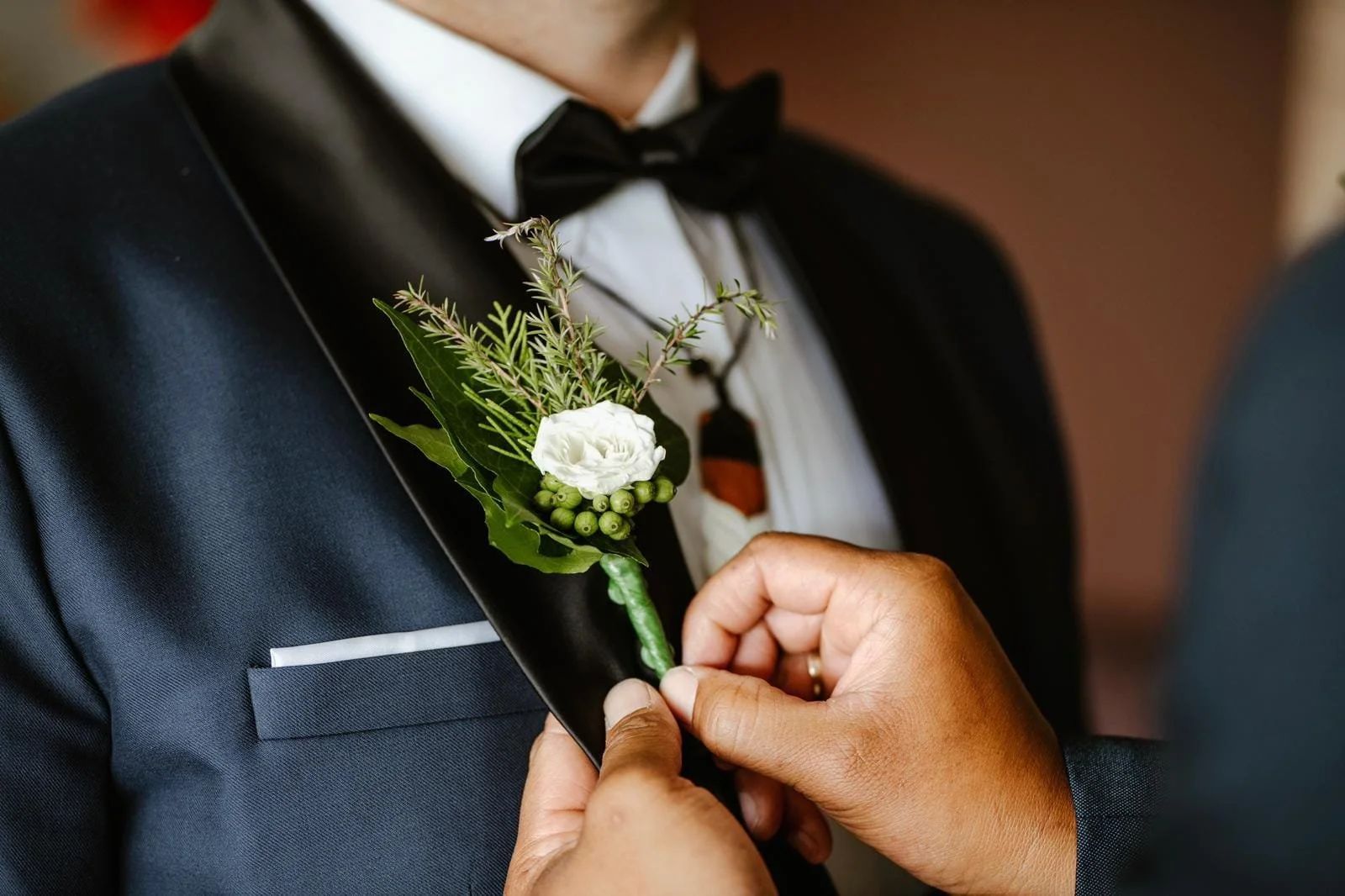 A person in a tuxedo has a boutonniere with a white flower, green leaves, and berries pinned to their lapel, as another individual adjusts it.