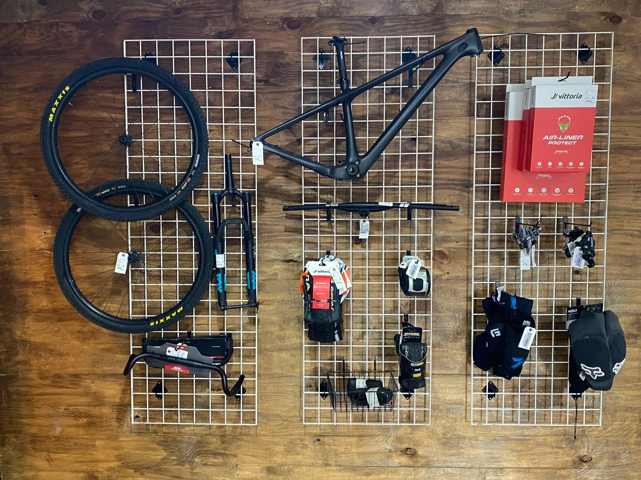 A wall display with a black mountain bike frame, two wheels, and various bike accessories and equipment arranged on a white metal grid against a wooden wall.