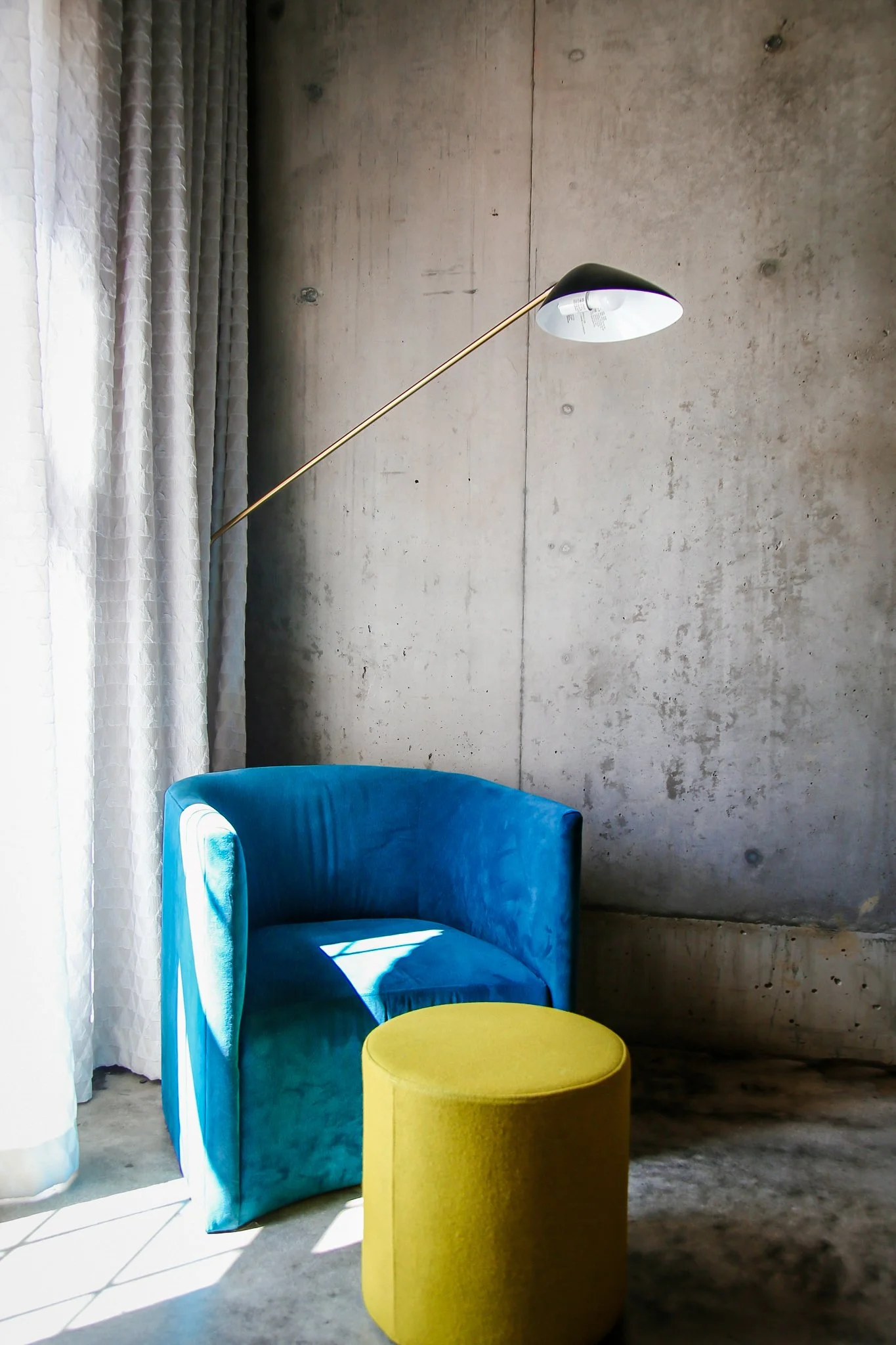 A modern blue velvet armchair with a yellow ottoman in front of a concrete wall, next to a window with white curtains, and a white and gold arc floor lamp.