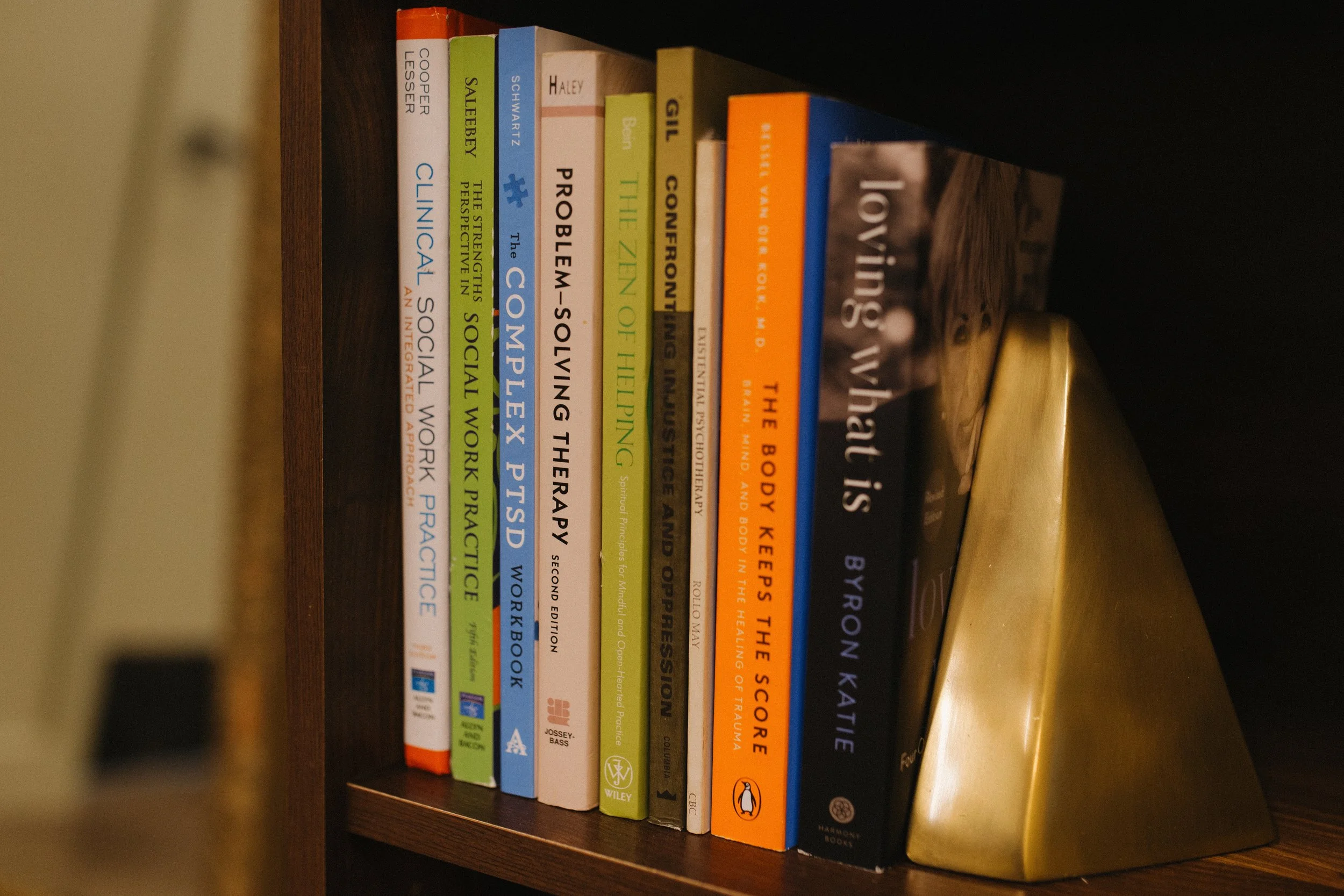 Books on a dark brown wooden shelf, including titles about therapy, social work, and psychology, with a large gold-colored decorative object beside them.