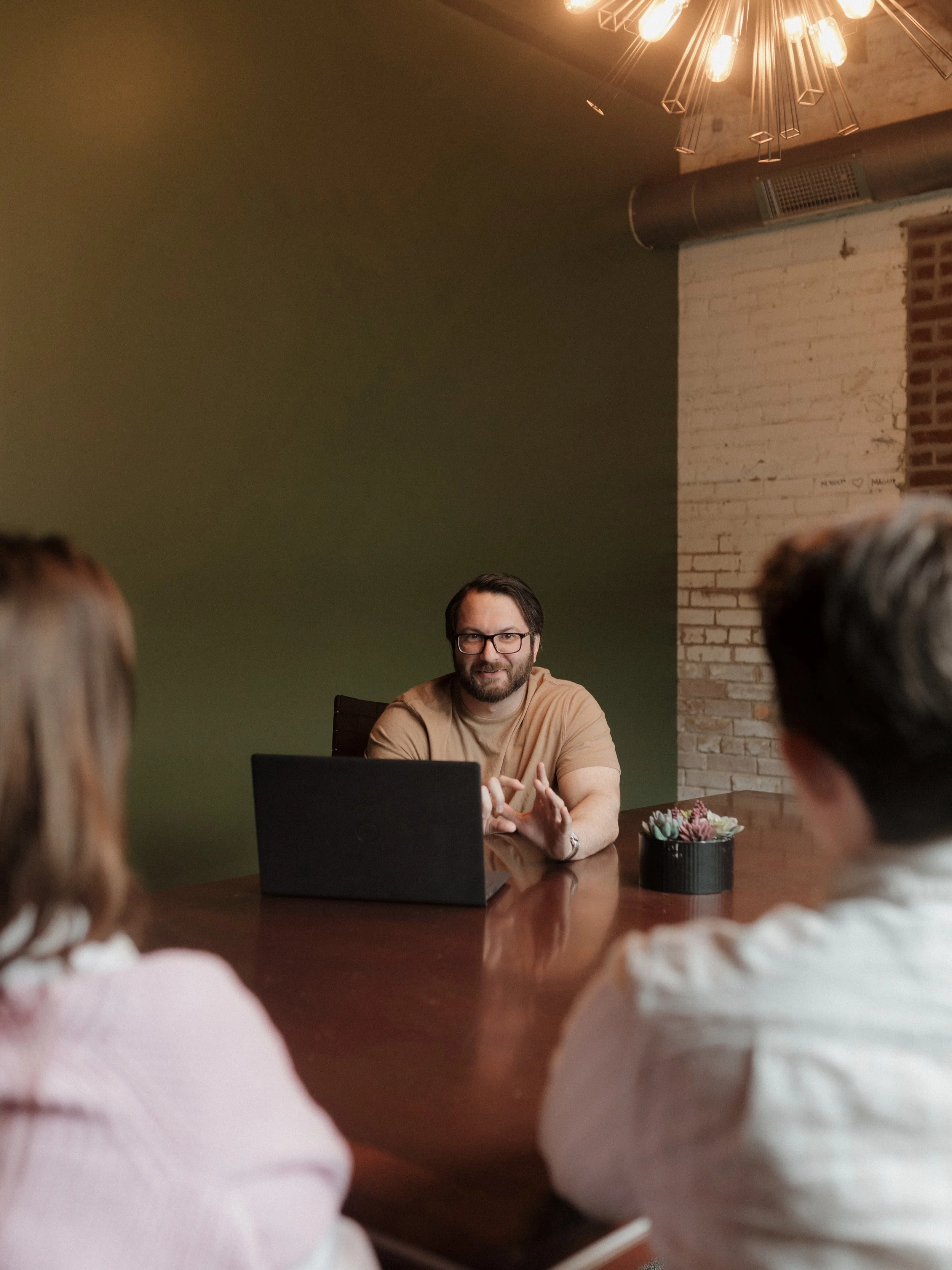 Man in glasses speaking to two people at a meeting table in a modern office.
