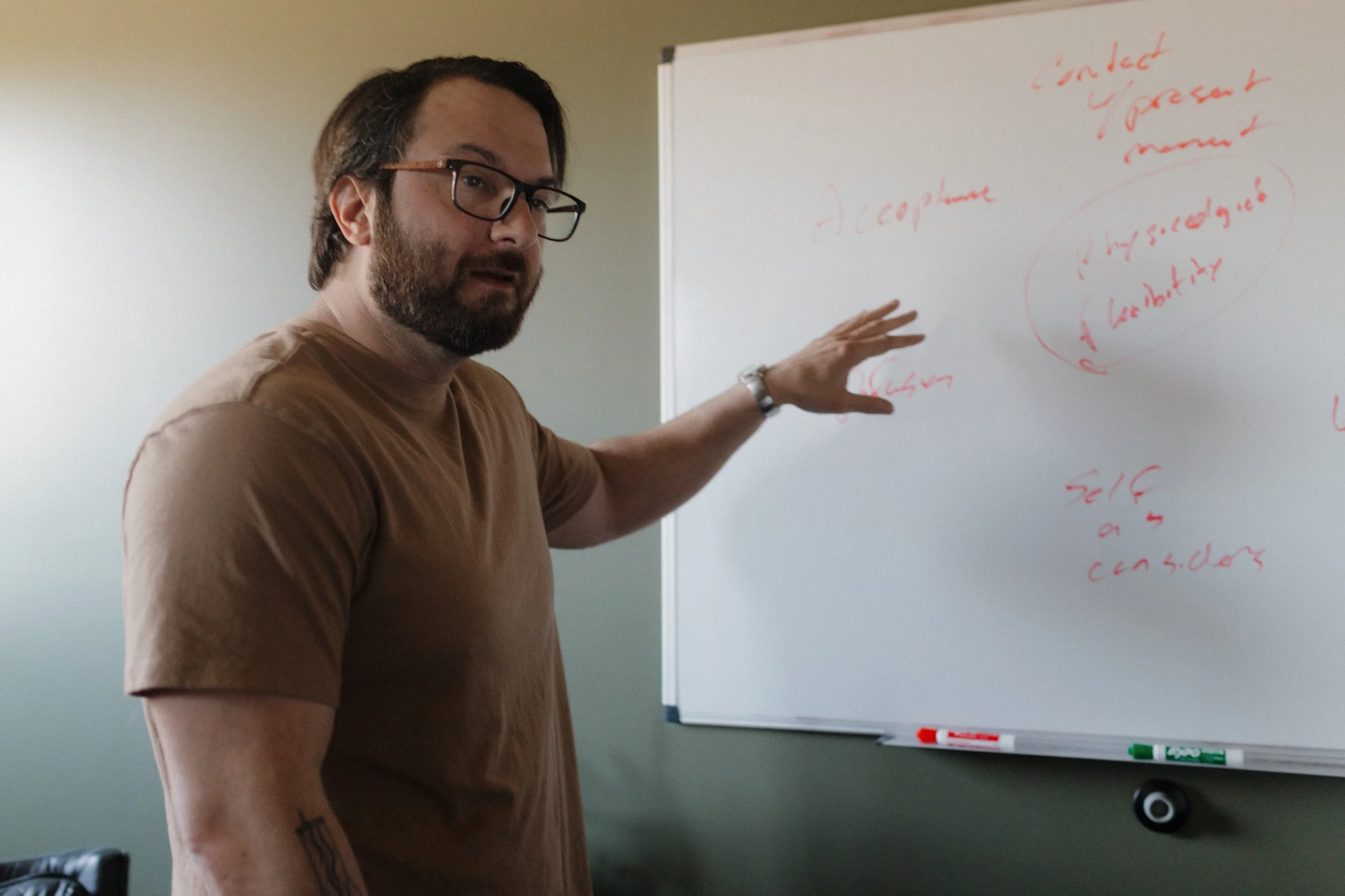 A man with glasses, a beard, and a tattoo on his arm pointing at a whiteboard with red handwritten notes.