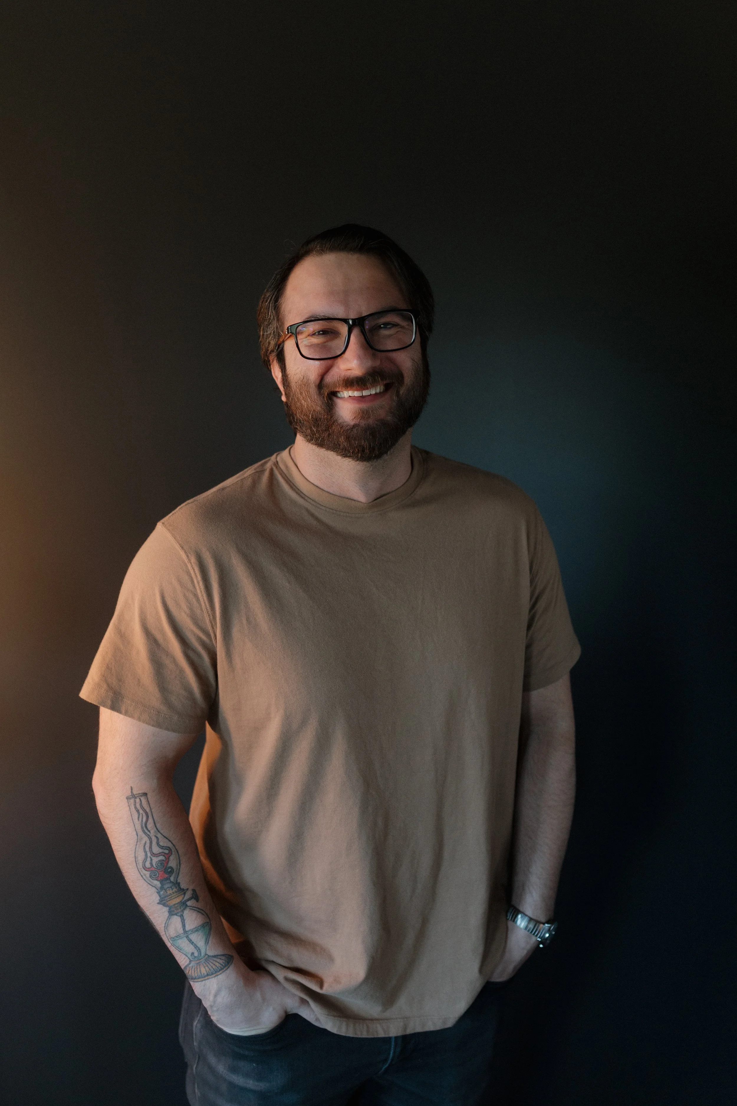 A man with glasses, a beard, and brown hair smiling, standing against a dark background with his hands in his pockets, wearing a beige t-shirt, jeans, and a wristwatch.