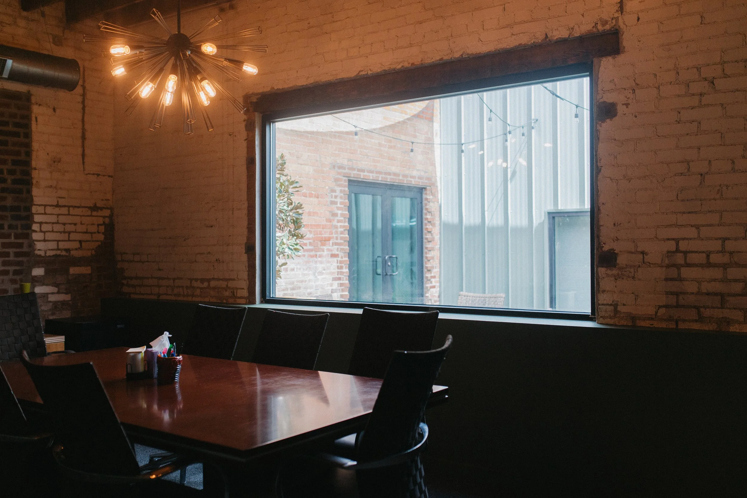Interior of a restaurant or conference room with a large window, brick walls, a wooden table, and black chairs, illuminated by a modern chandelier with multiple exposed bulbs.