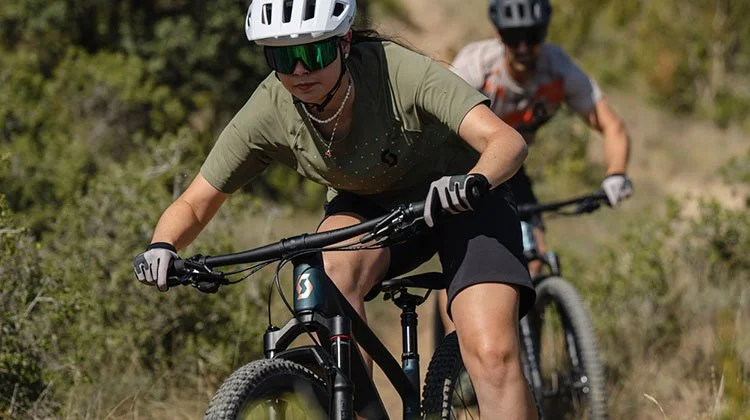 Two mountain bikers riding on a trail with greenery, wearing helmets and sportswear.