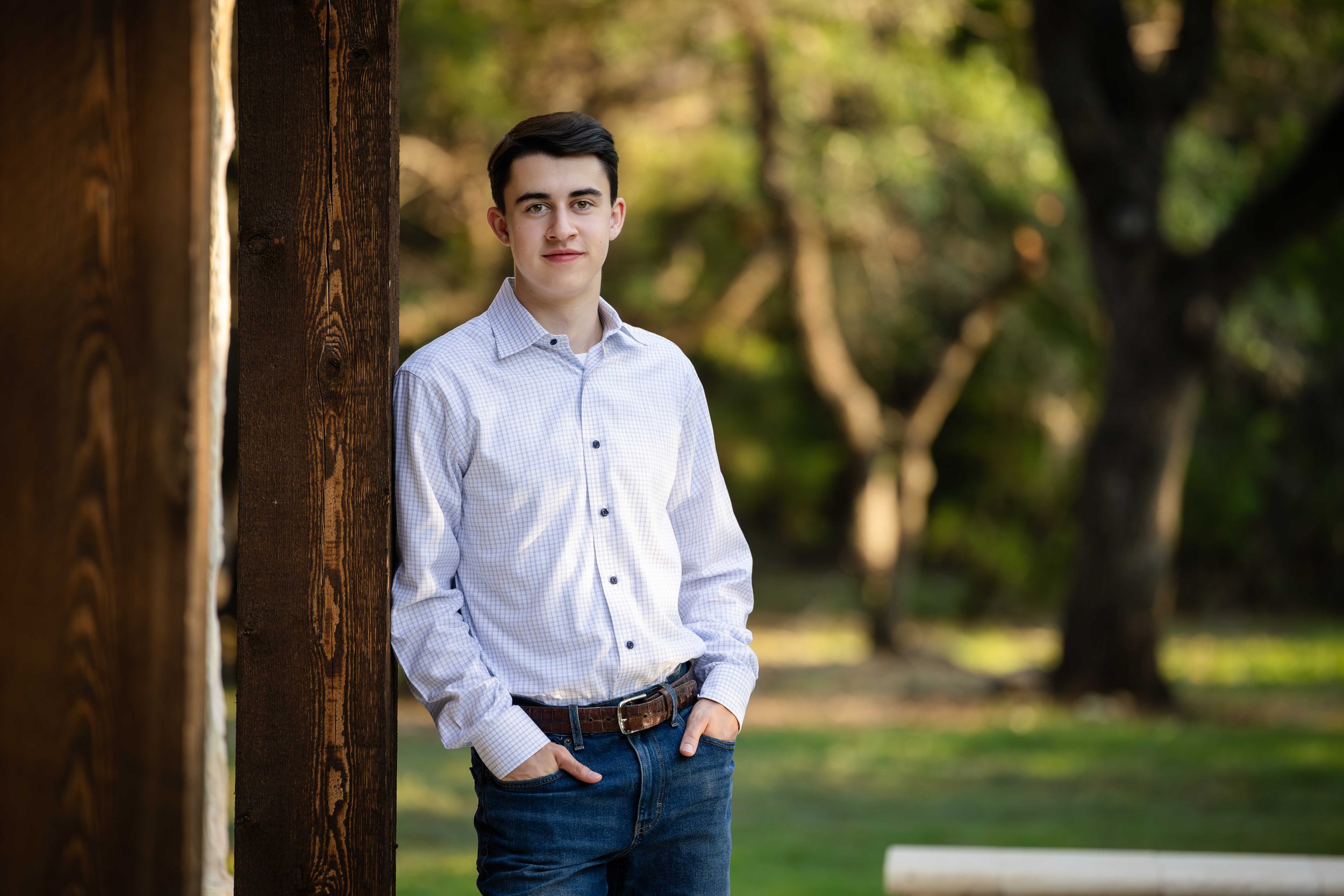 Young man with dark hair wearing a white checkered shirt and blue jeans, standing outdoors near a wooden post, with trees and greenery in the background.