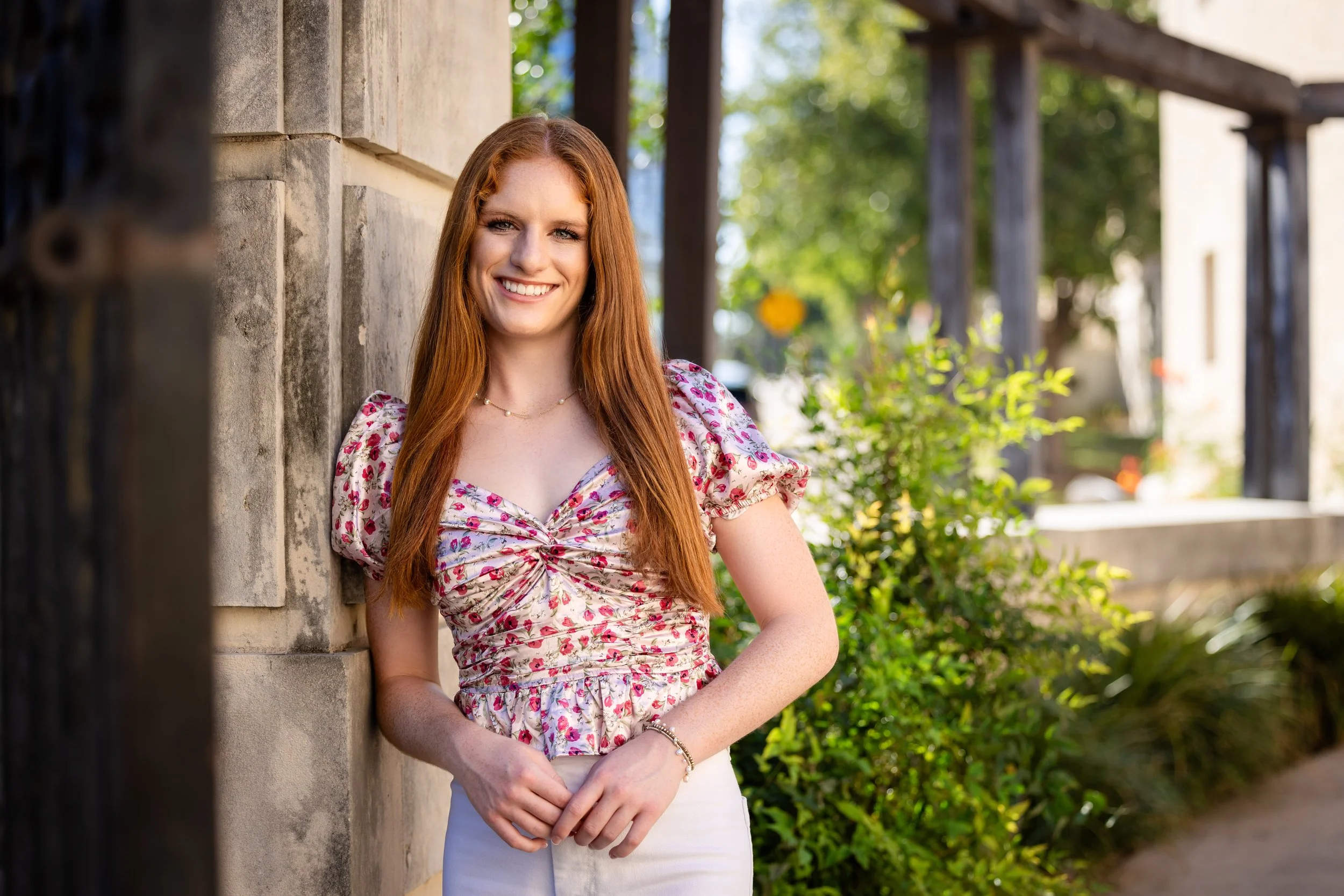 A young woman with long red hair, wearing a floral top and white pants, is smiling and standing outdoors next to a stone wall with greenery in the background.