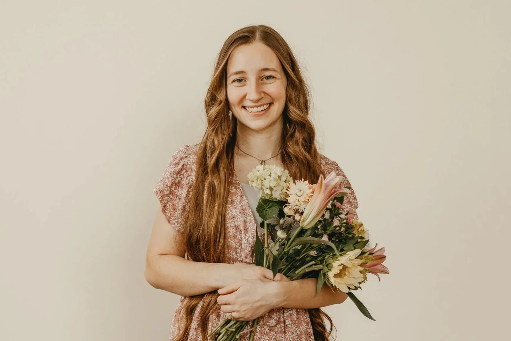 Young woman with long red hair smiling while holding a bouquet of various flowers in front of a plain light-colored wall.