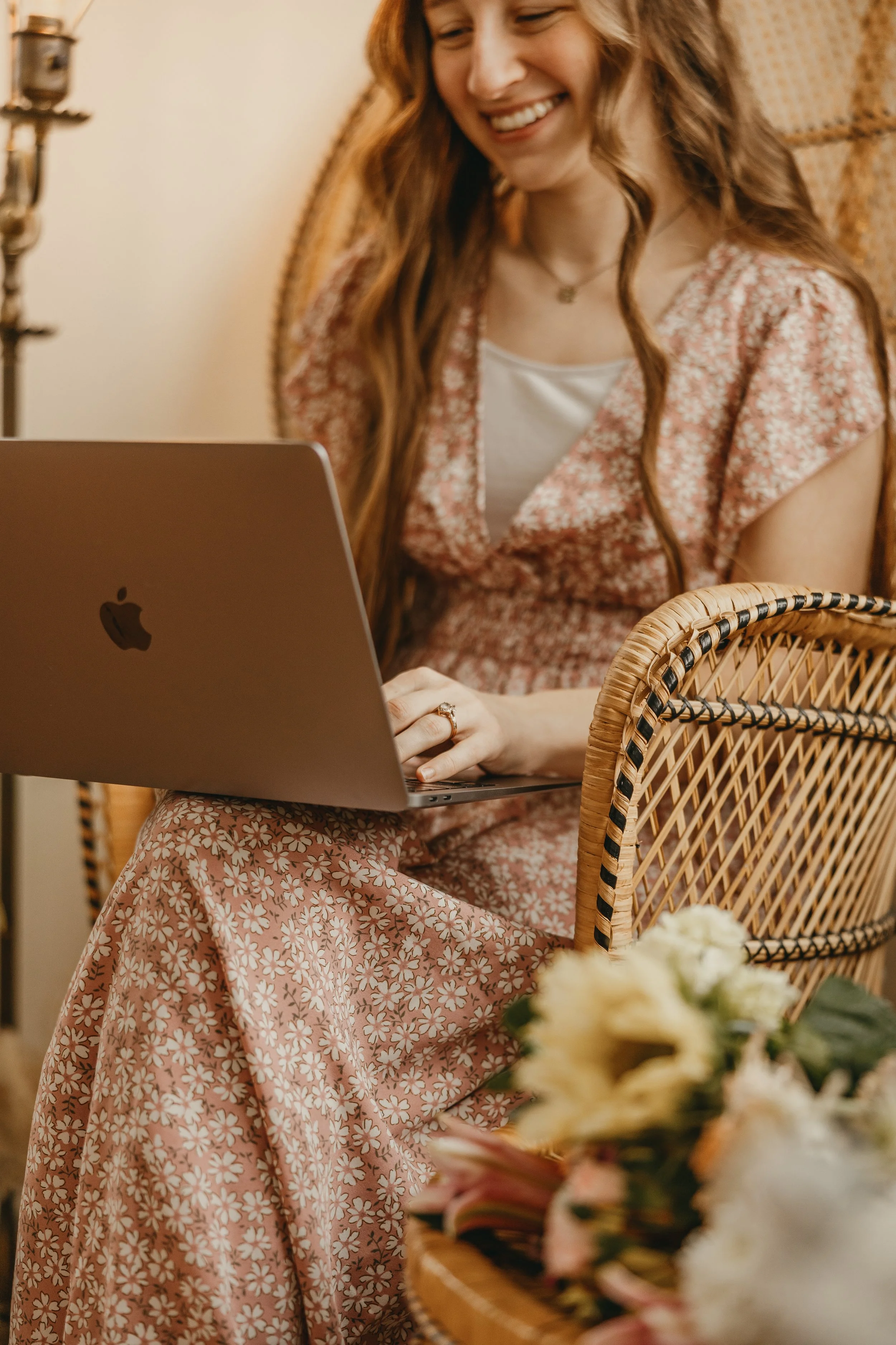 A smiling woman with long wavy hair, wearing a floral dress, using a silver MacBook laptop, seated in a wicker chair with a bouquet of flowers in the foreground.
