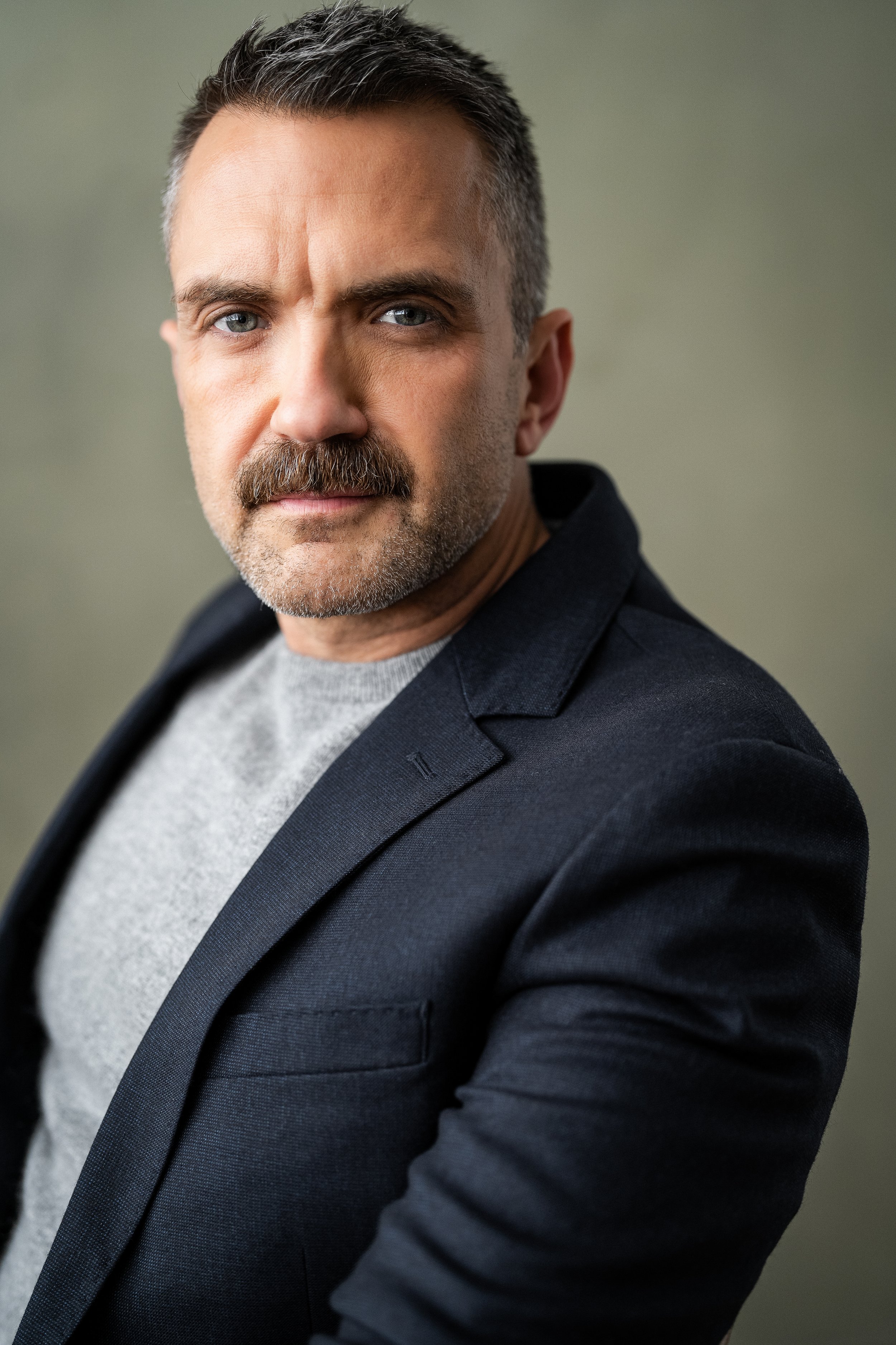 Close-up of a middle-aged man with short dark hair, a beard, wearing a dark blazer and a light gray T-shirt, posed against a plain background.