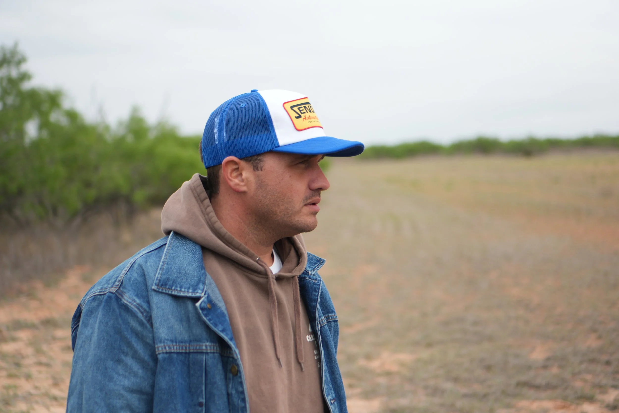 A man wearing a blue mesh trucker hat with a yellow and red patch, a tan hoodie, and a blue denim jacket stands outdoors in a rural area with green trees and an overcast sky in the background.