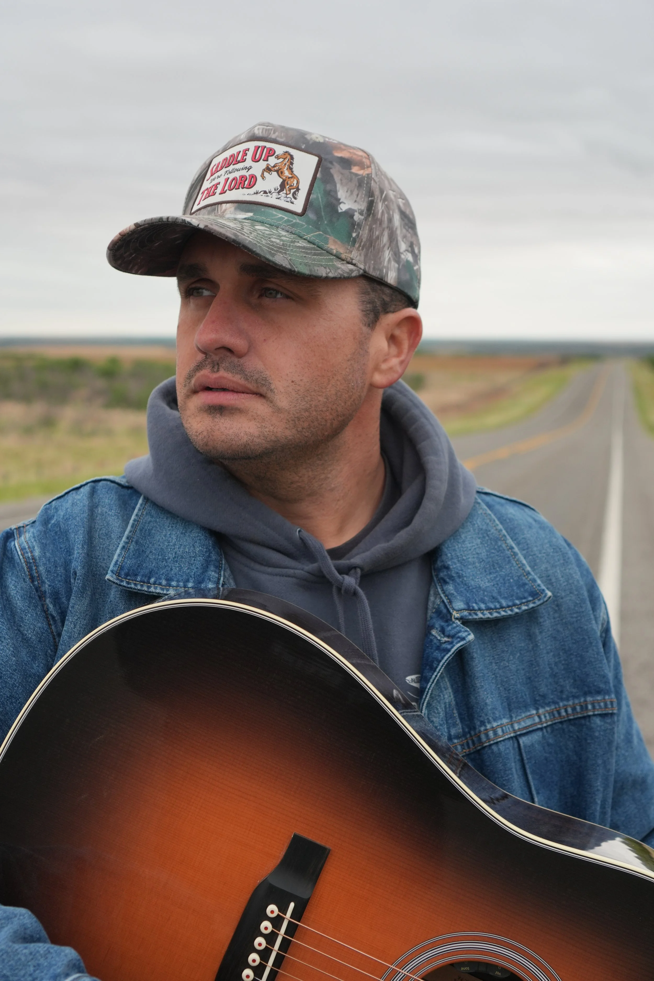 A man wearing a camouflage baseball cap with the words 'Saddle Up the Lord' and a horse logo, along with a denim jacket and hoodie, holding an acoustic guitar outdoors on a cloudy day with a rural road in the background.