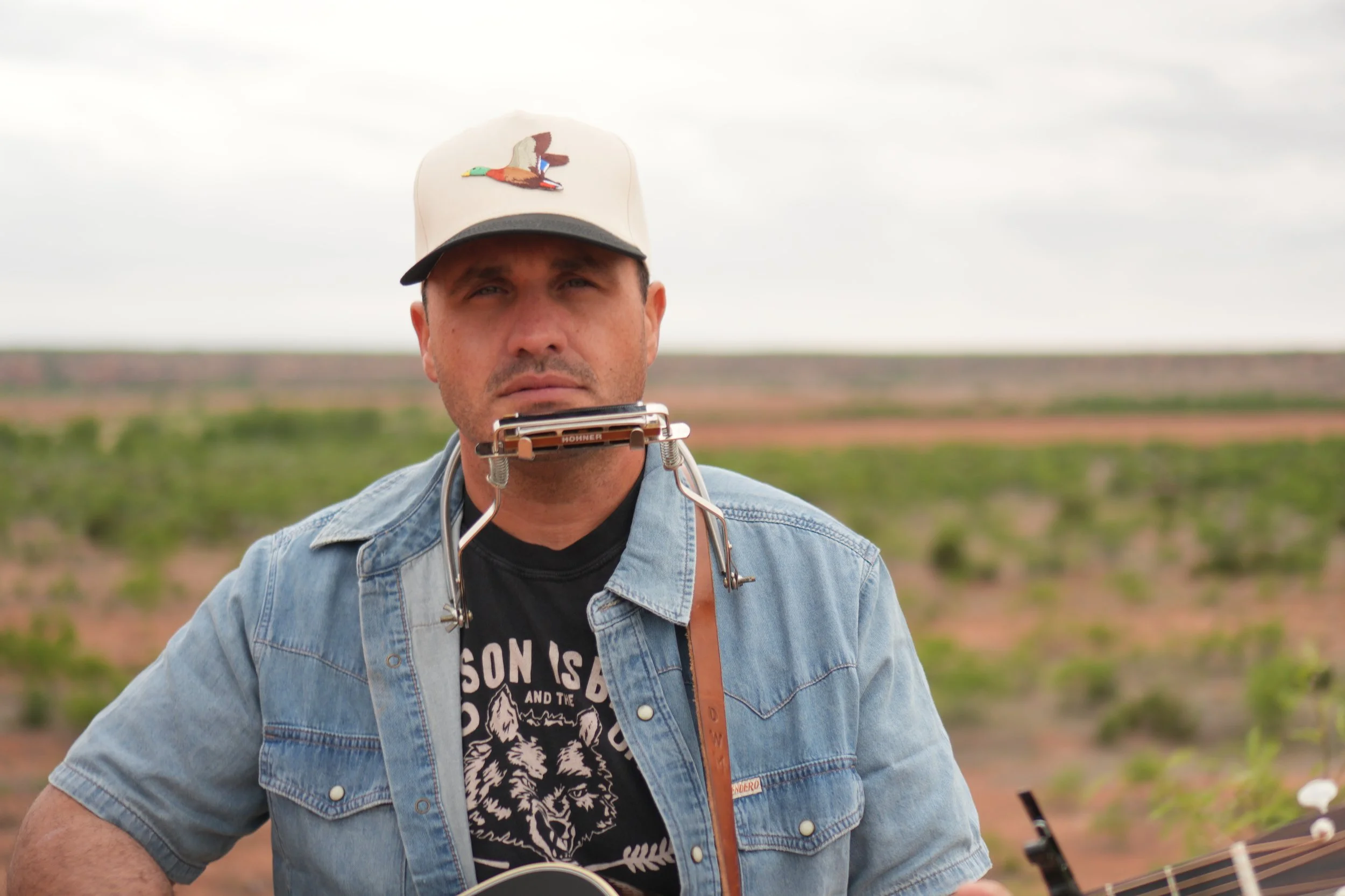 A man stands outdoors in a field, playing a harmonica and holding a guitar, wearing a beige baseball cap with a colorful duck embroidery and a denim jacket.