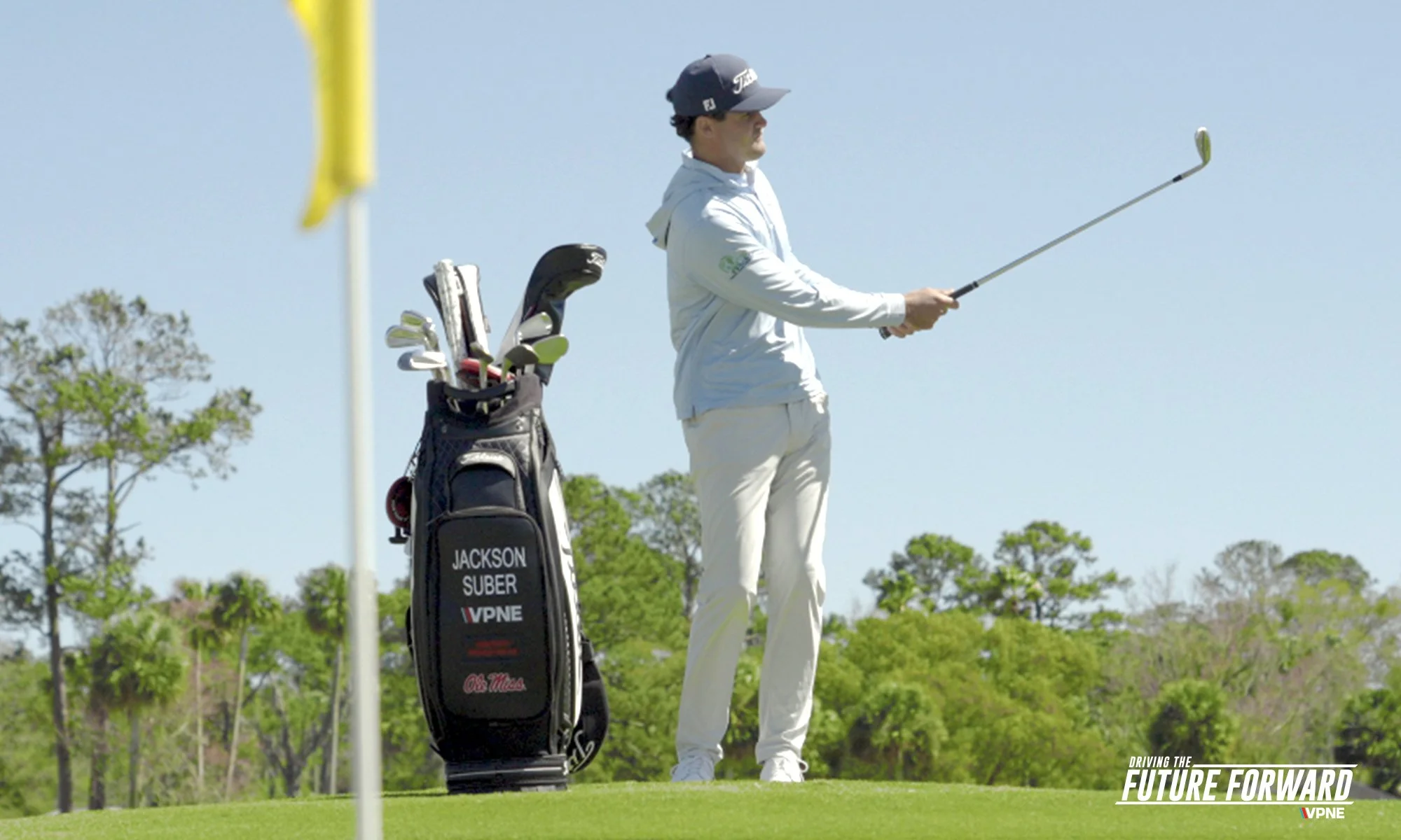 A golfer stands on a golf course, holding a golf club, with a golf bag beside him. The course is lush and green, with trees in the background, and a clear sky above. The image features a logo in the bottom right that says "Driving the Future Forward" with the VPNE logo underneath.
