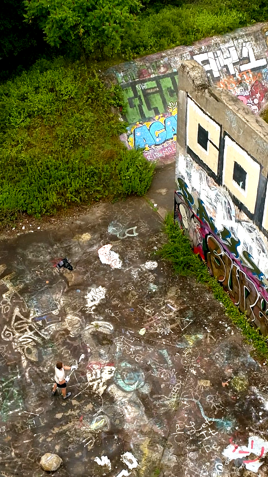 Graffiti-covered concrete ground with a person holding a paint roller. There are two people in the image. The scene is in an outdoor area surrounded by graffiti on walls and green bushes.
