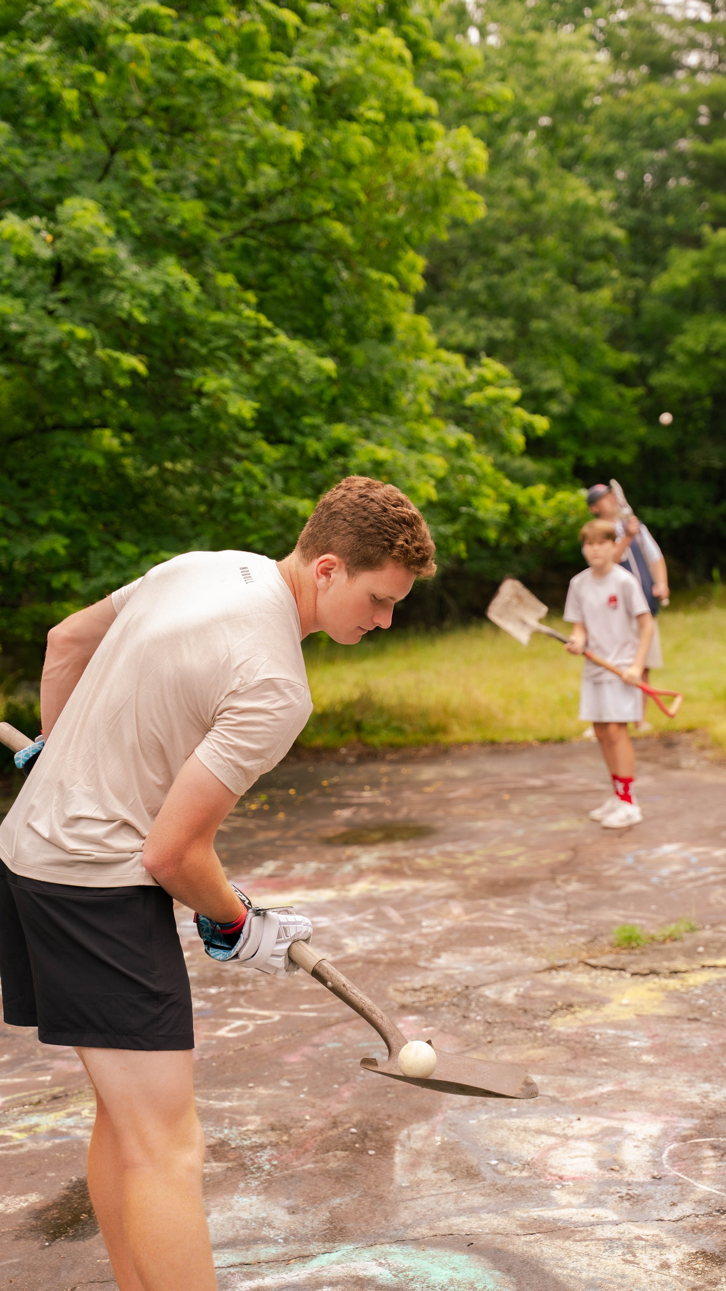 A young man is playing lacrosse outdoors, holding a stick and playing wall ball at a paved court surrounded by green trees, with children in the background holding shovels.