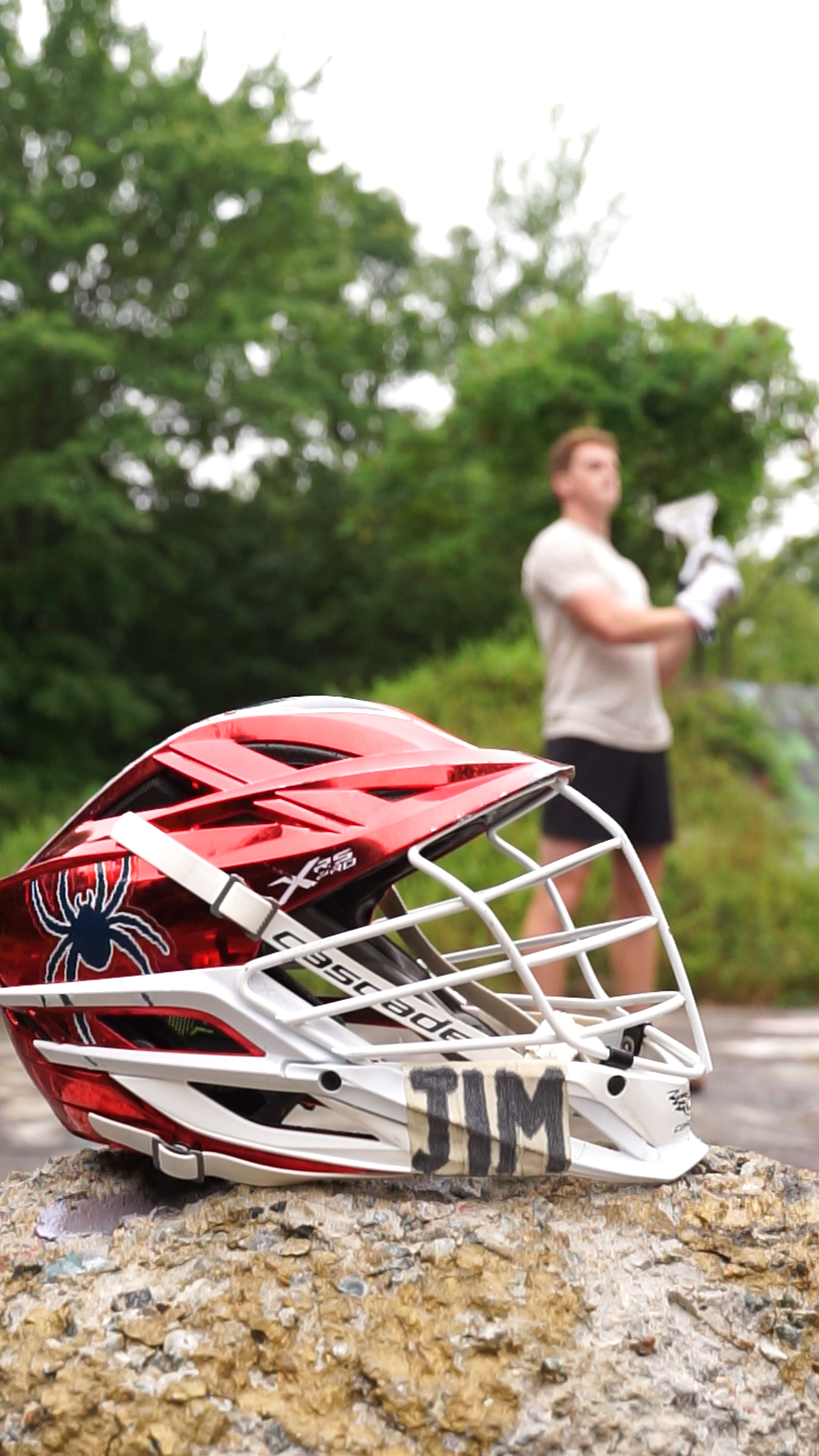 A red and white lacrosse helmet with a spider logo on the side is resting on a rock. In the background, a woman is standing on a trail holding a lacrosse stick, with trees and greenery around her.