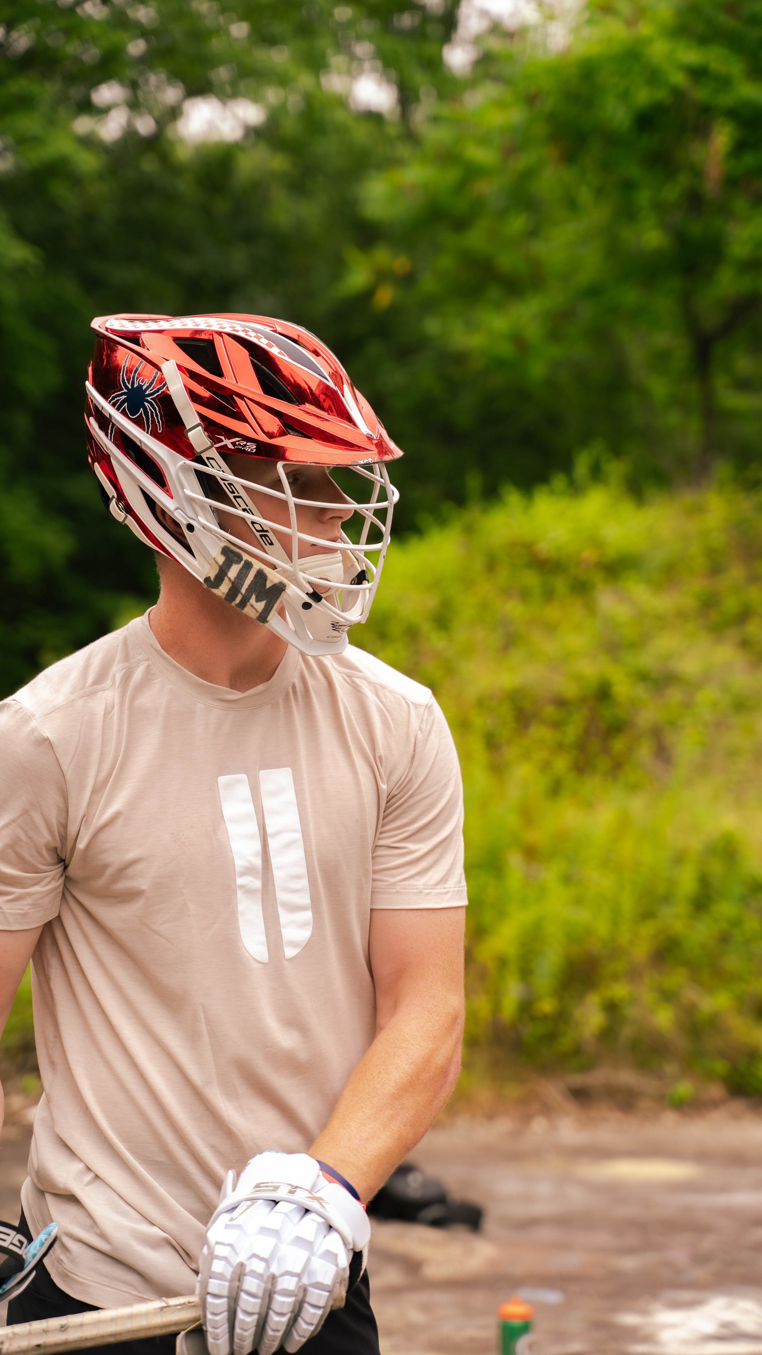 A person wearing a red and white helmet, beige t-shirt, and white gloves, standing outdoors with a green wooded background.