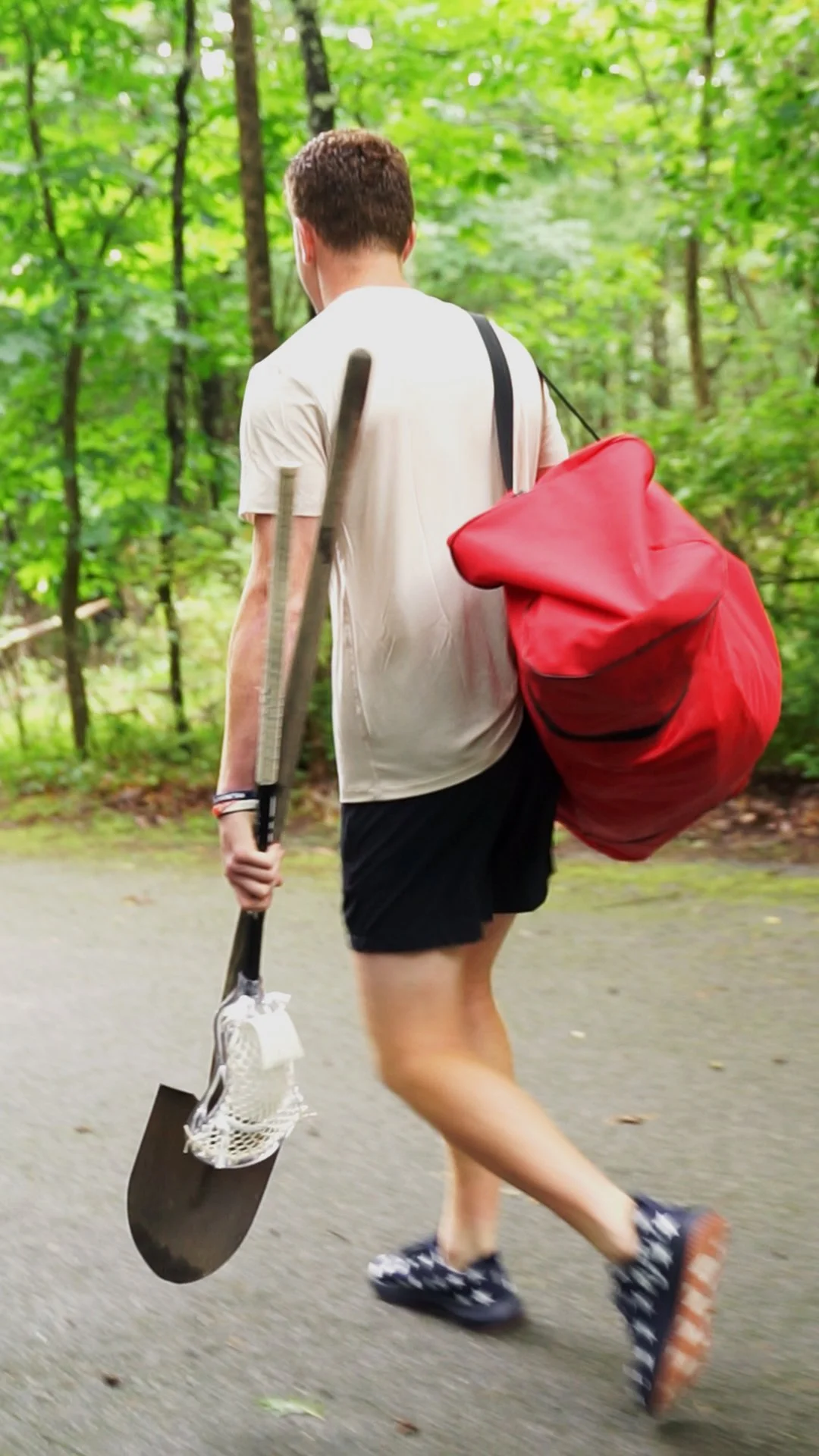 A person with short hair walking in a wooded area, carrying a large red bag on their shoulder and holding a shovel.