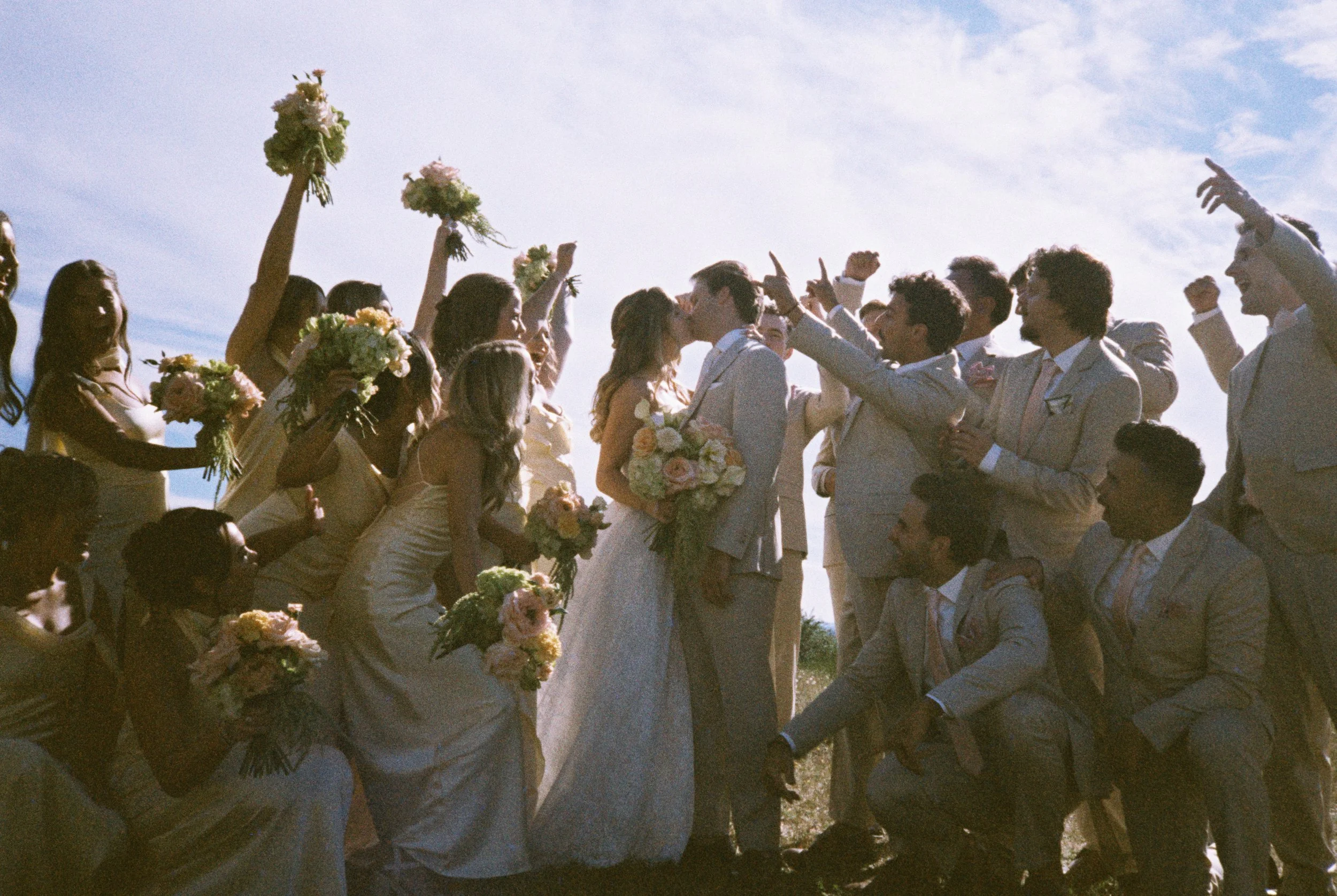 bride and groom with bridal party kissing at sunset.