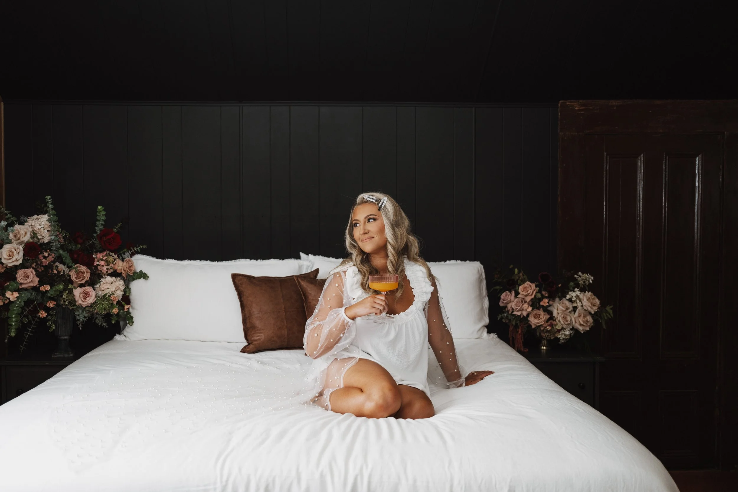 bride getting ready on a white bed flanked by moody burgundy and mauve floral arrangements with a cocktail in hand.