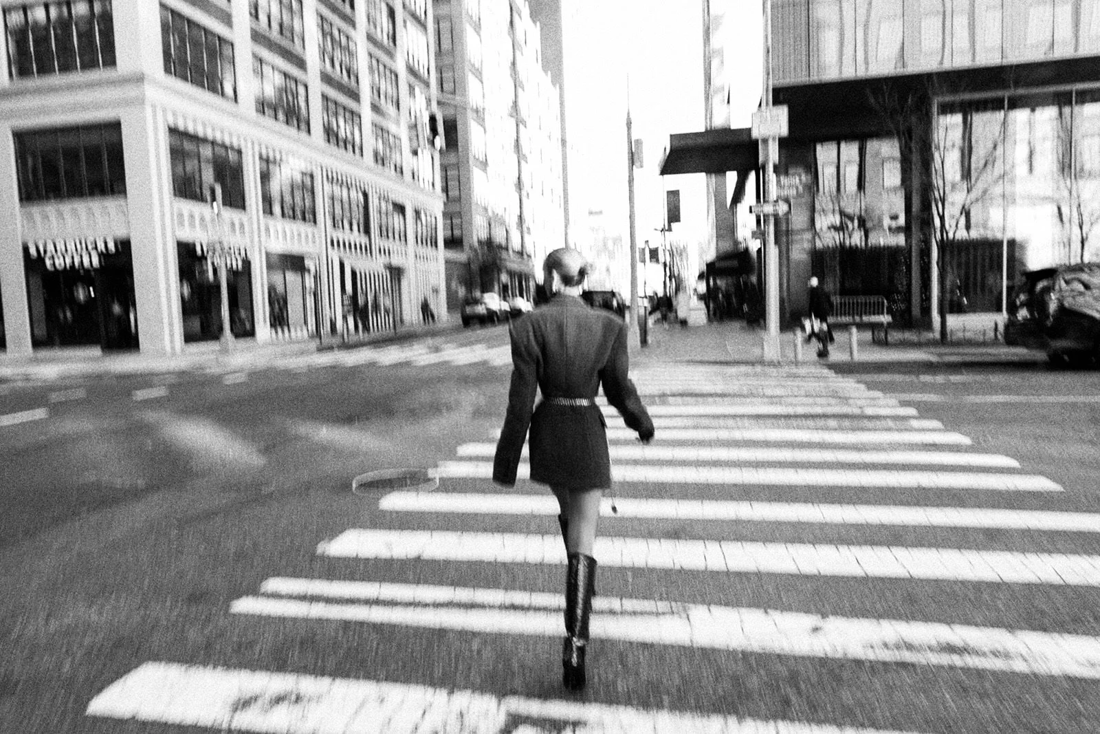 Black and white photo of Virginia Bundy in a belted blazer and high boots walking across a city crosswalk, with tall buildings and street scenes of New York City in the background.